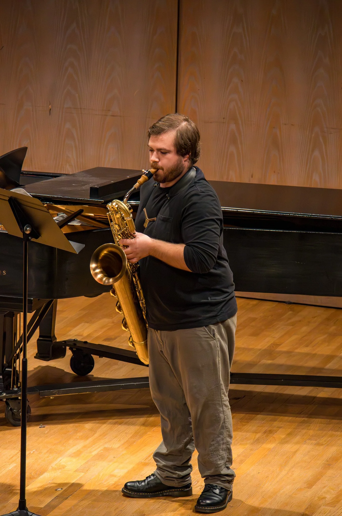 A man playing a saxophone in a music performance space, standing on a wooden floor with a piano in the background and a music stand in front.
