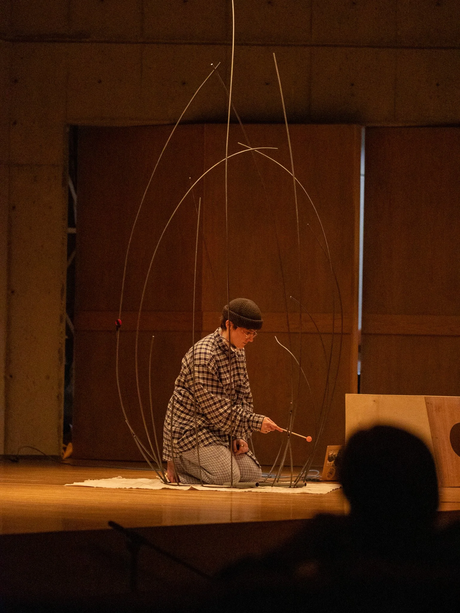 A person kneeling on stage, wearing a plaid shirt, pants, and a beanie, surrounded by thin, curved metal rods forming a sculpture or artistic installation.
