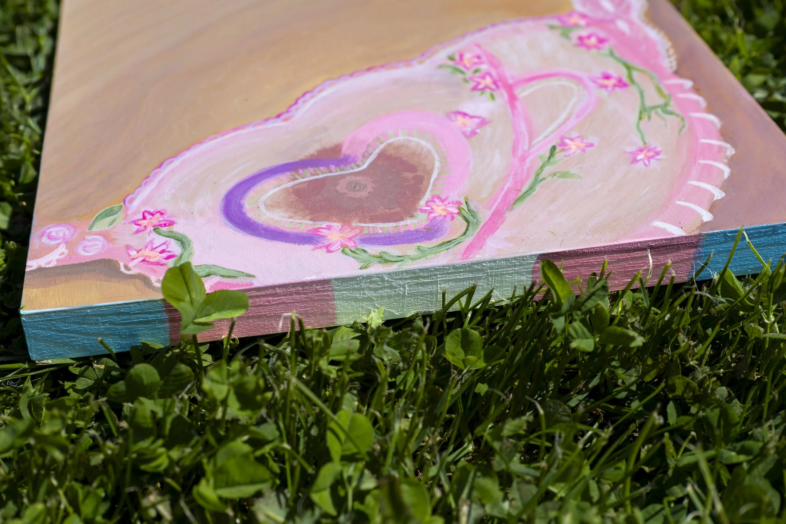 A painted canvas with pink hearts, flowers, and decorative designs lying on green grass.