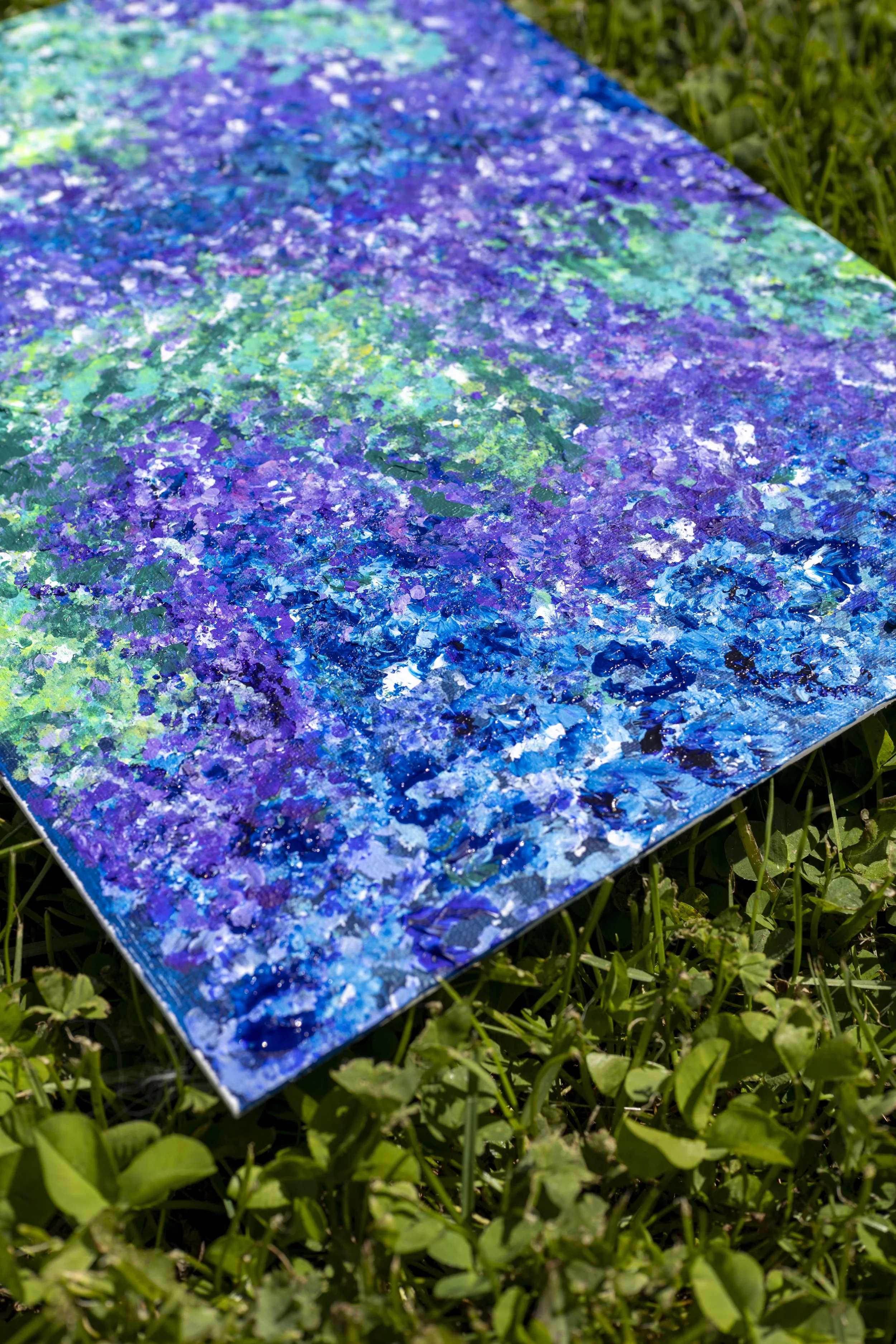 An abstract painting with a mix of purple, blue, green, and white colors lying on green grass.
