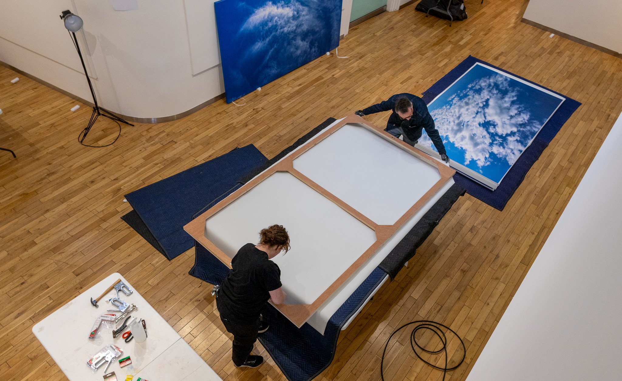 People assembling large printed images of the sky with clouds on frames in an indoor space, possibly an art gallery or exhibition area.