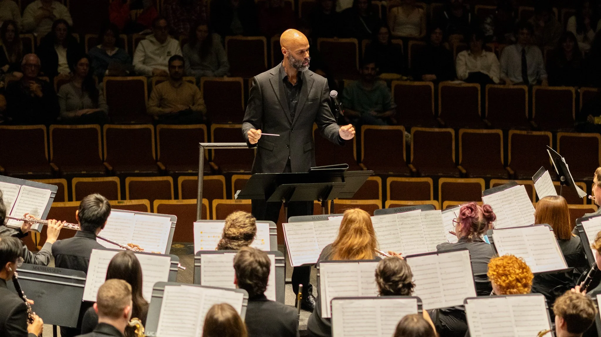 A man in a black suit stands at a conductor's podium during a symphony orchestra performance, with musicians and sheet music in front and an audience in the background.