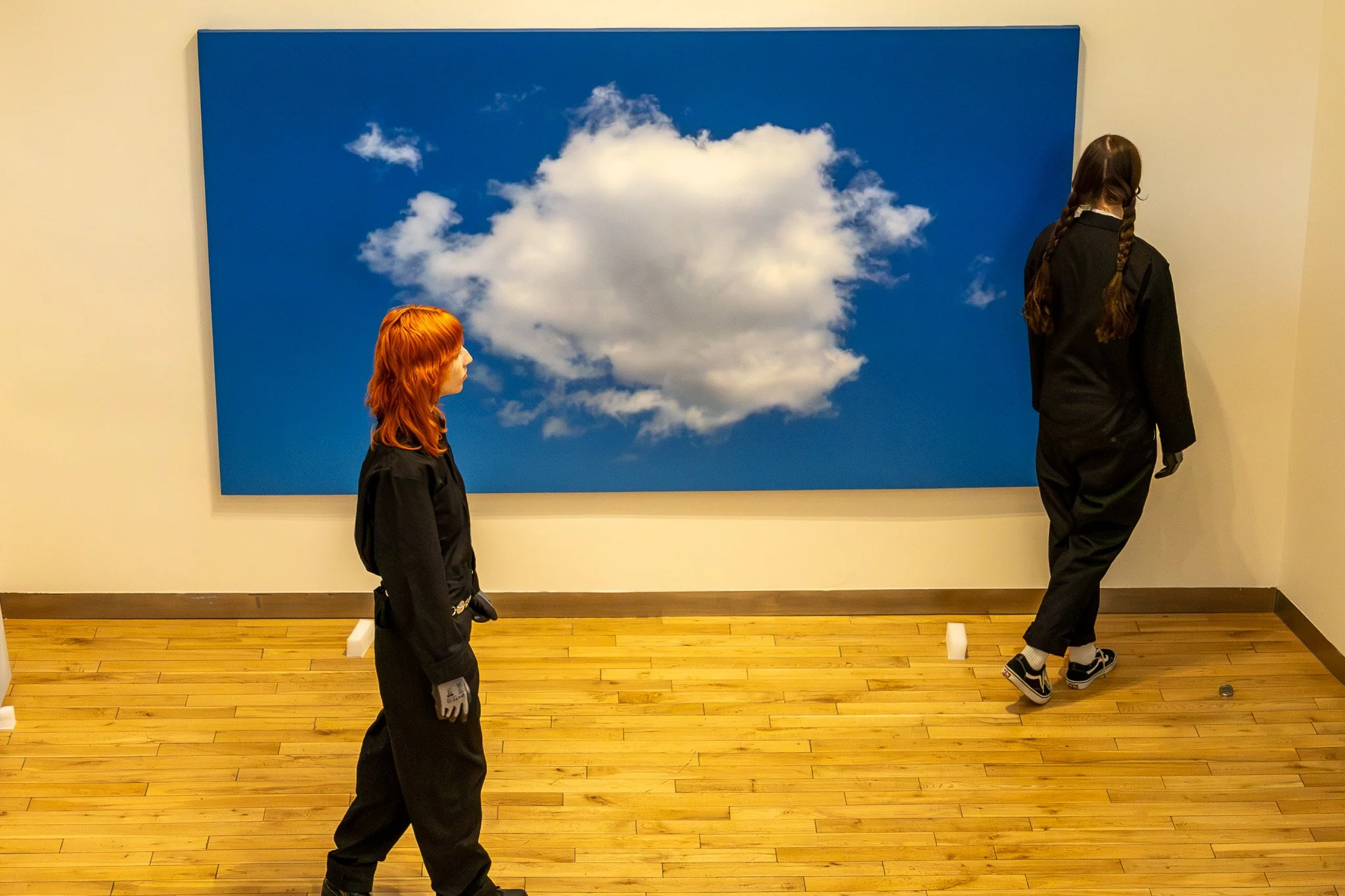 Two girls in black outfits standing in front of a large blue painting of a white cloud in the sky, one girl with ginger hair and the other with long braid and dark hair.