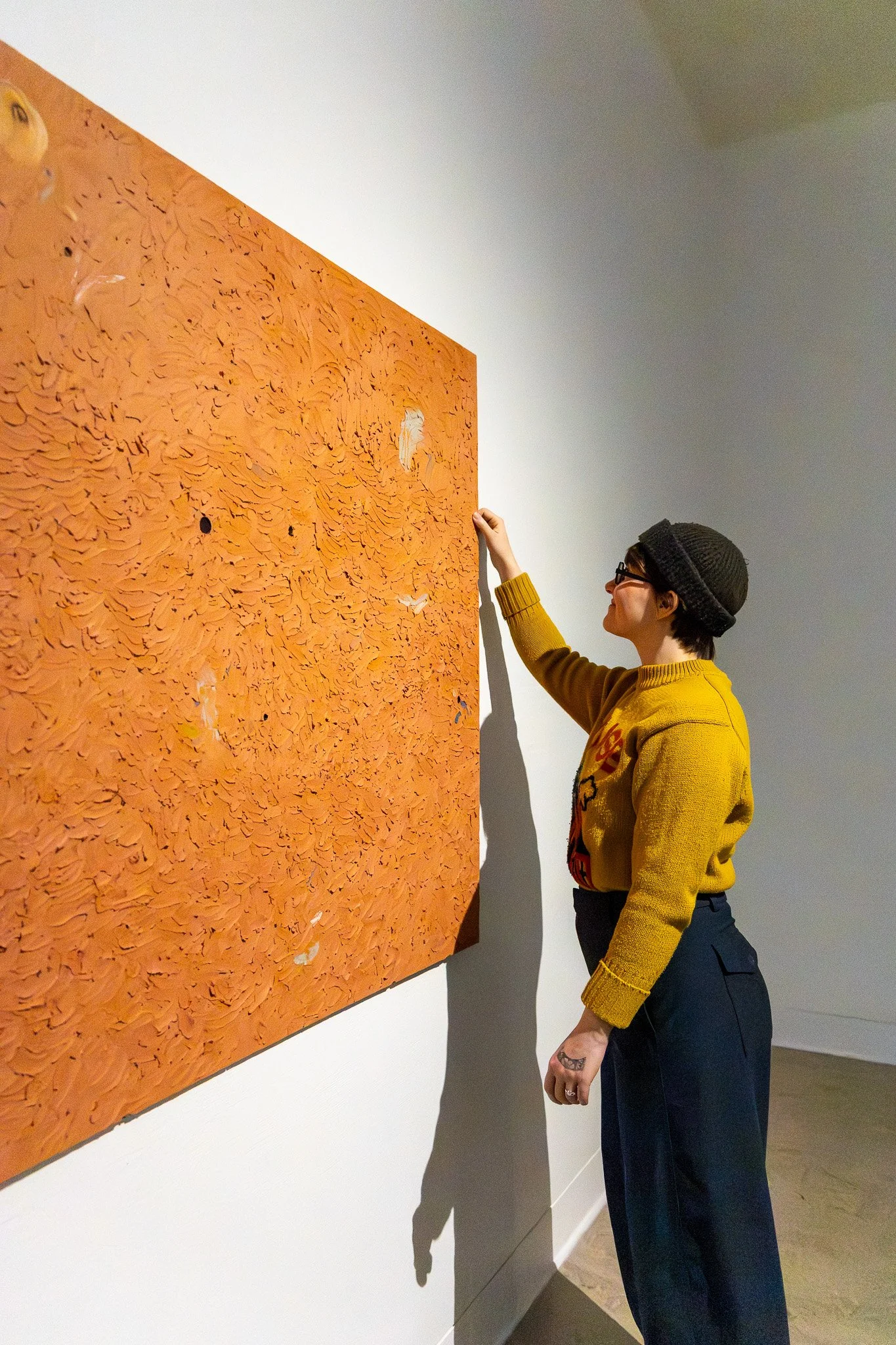 A woman with short hair, glasses, a black knit beret, and a yellow sweater is standing in an art gallery, looking at and gently touching a textured orange artwork on the wall.