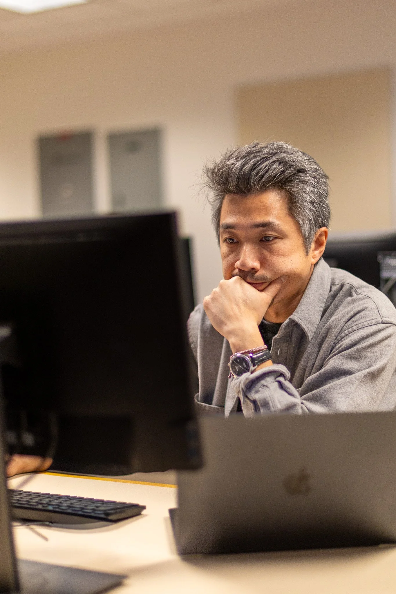 A man with gray hair and a gray shirt sitting at a desk, looking at a computer monitor with thoughtful expression, in an office setting.