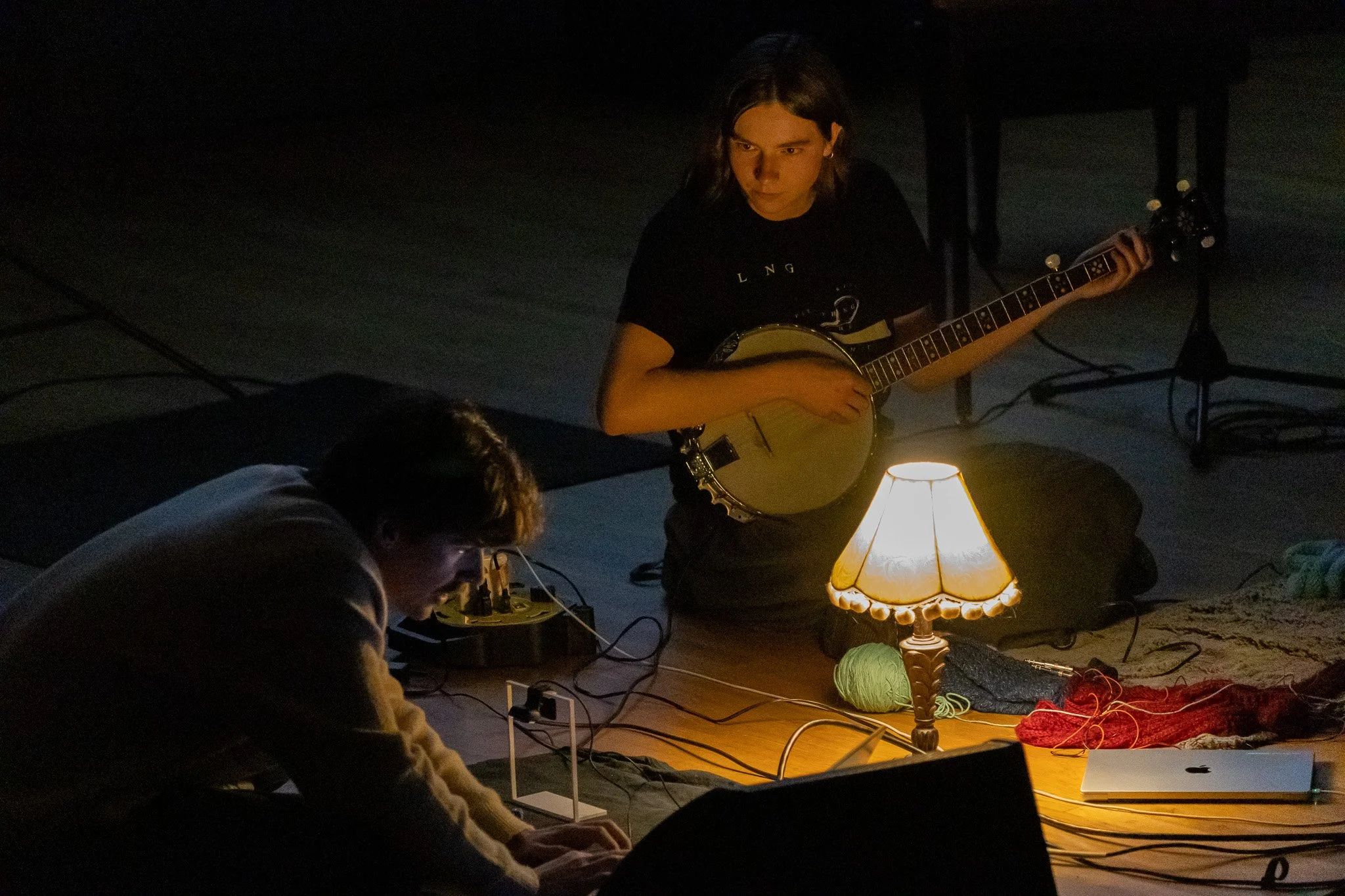 A young woman playing an acoustic guitar in a dimly lit room illuminated by a table lamp, with yarn and knitting supplies around her, and a person sitting nearby.