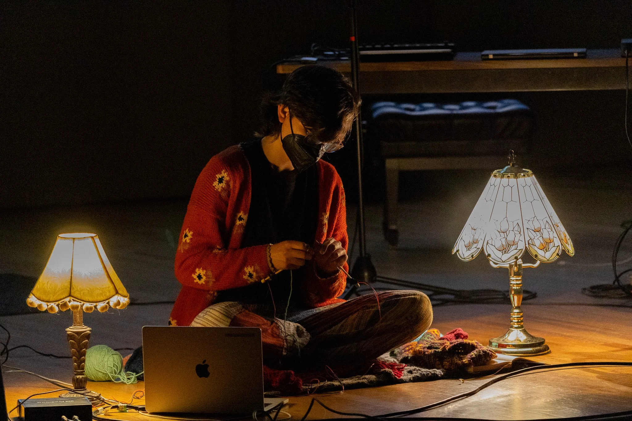 Person sitting cross-legged on the floor in a dark room, wearing a red sweater with yellow patterns, black face mask, and glasses, working with knitting needles. Two decorative lamps provide warm lighting on either side, and yarn and a laptop are pla