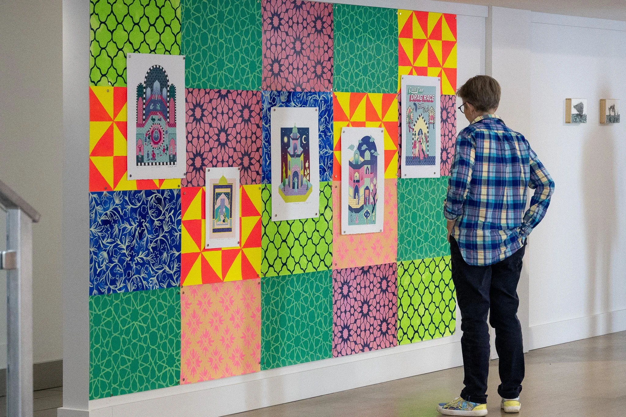 A person wearing a colorful plaid shirt and dark pants stands in front of a display of vibrant, patterned fabric or artwork on a wall, with various framed illustrations or posters hanging among the textiles.