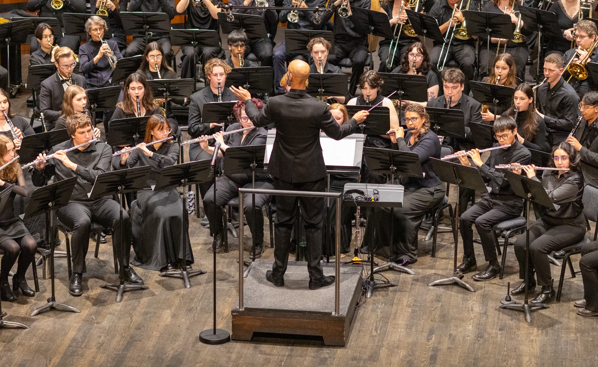 Orchestra performing on stage with the conductor in front, members playing various instruments including flutes, clarinets, saxophones, trumpets, and trombones.