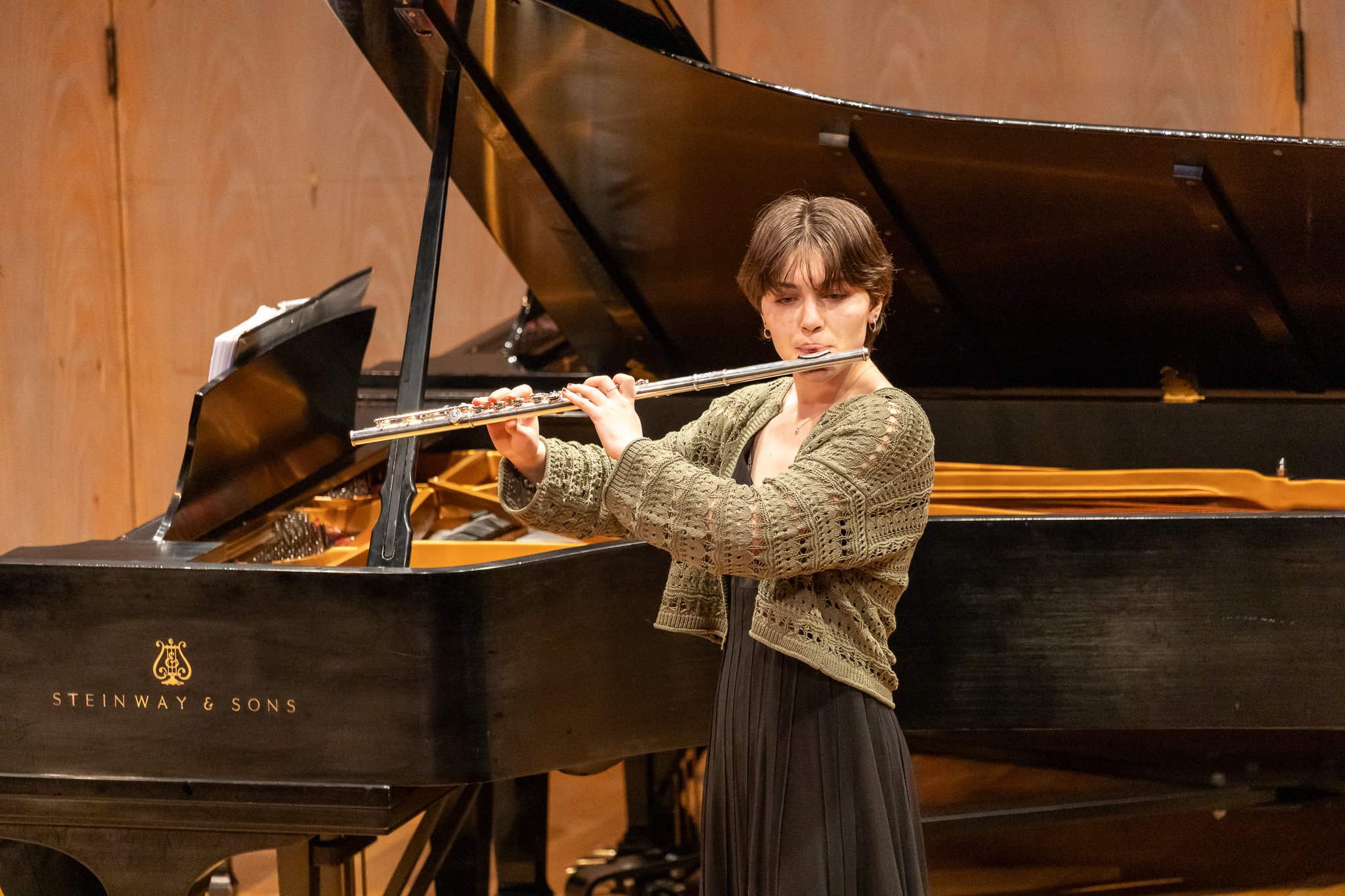 A young woman playing a flute on stage with a grand piano in the background.