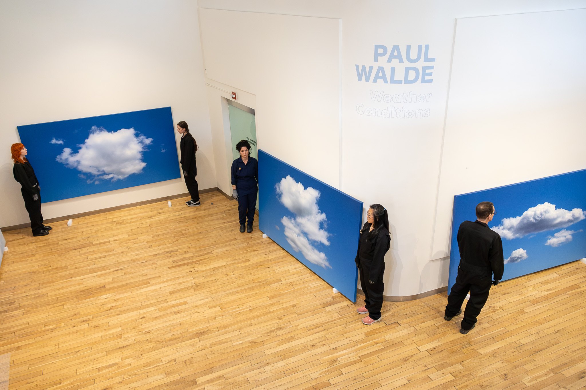 Gallery wall with large photos of cloudy blue skies, with several people observing the artwork.