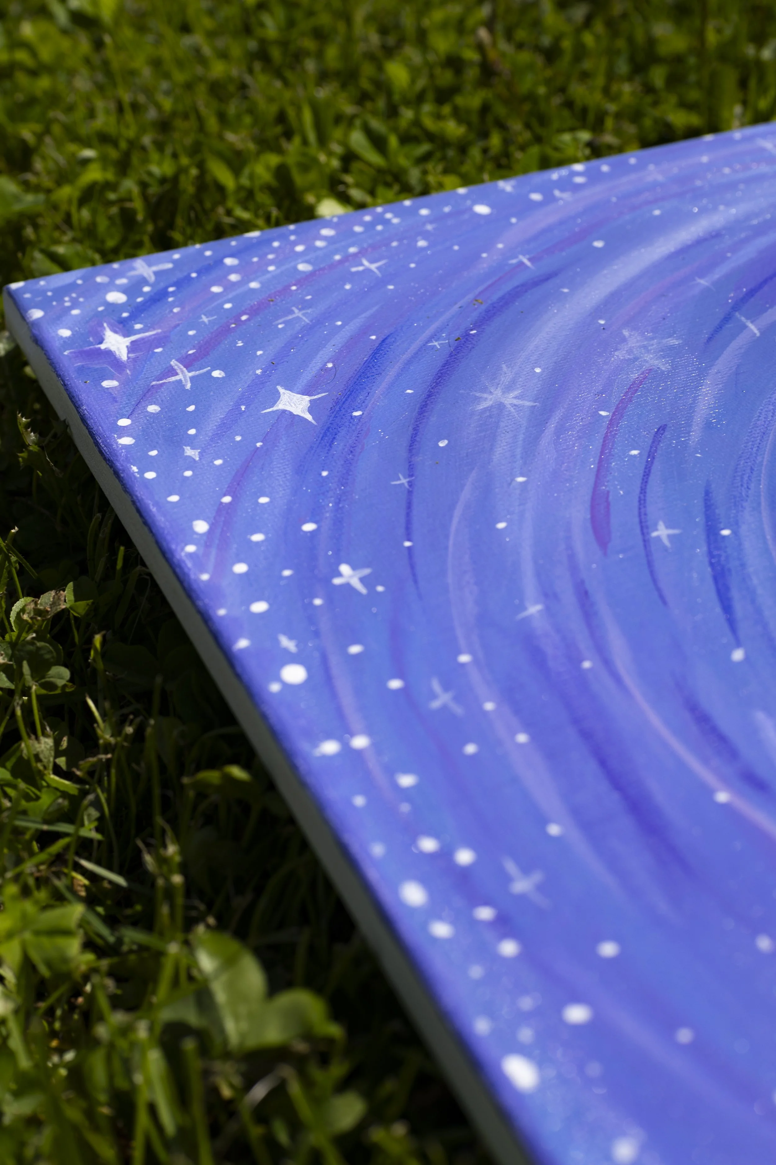 Close-up of a painted canvas with a galaxy scene, featuring blue and purple swirls, white stars, and dots, placed on grass.