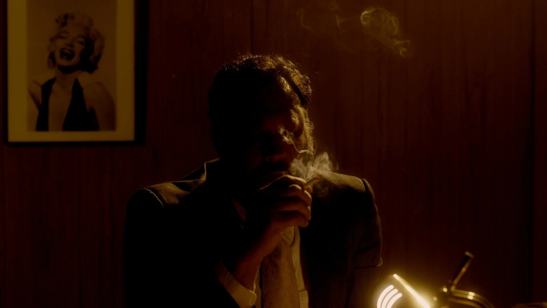 A man with dark curly hair sitting in dim lighting, smoking a  cigarette, with a smoke ring rising.
