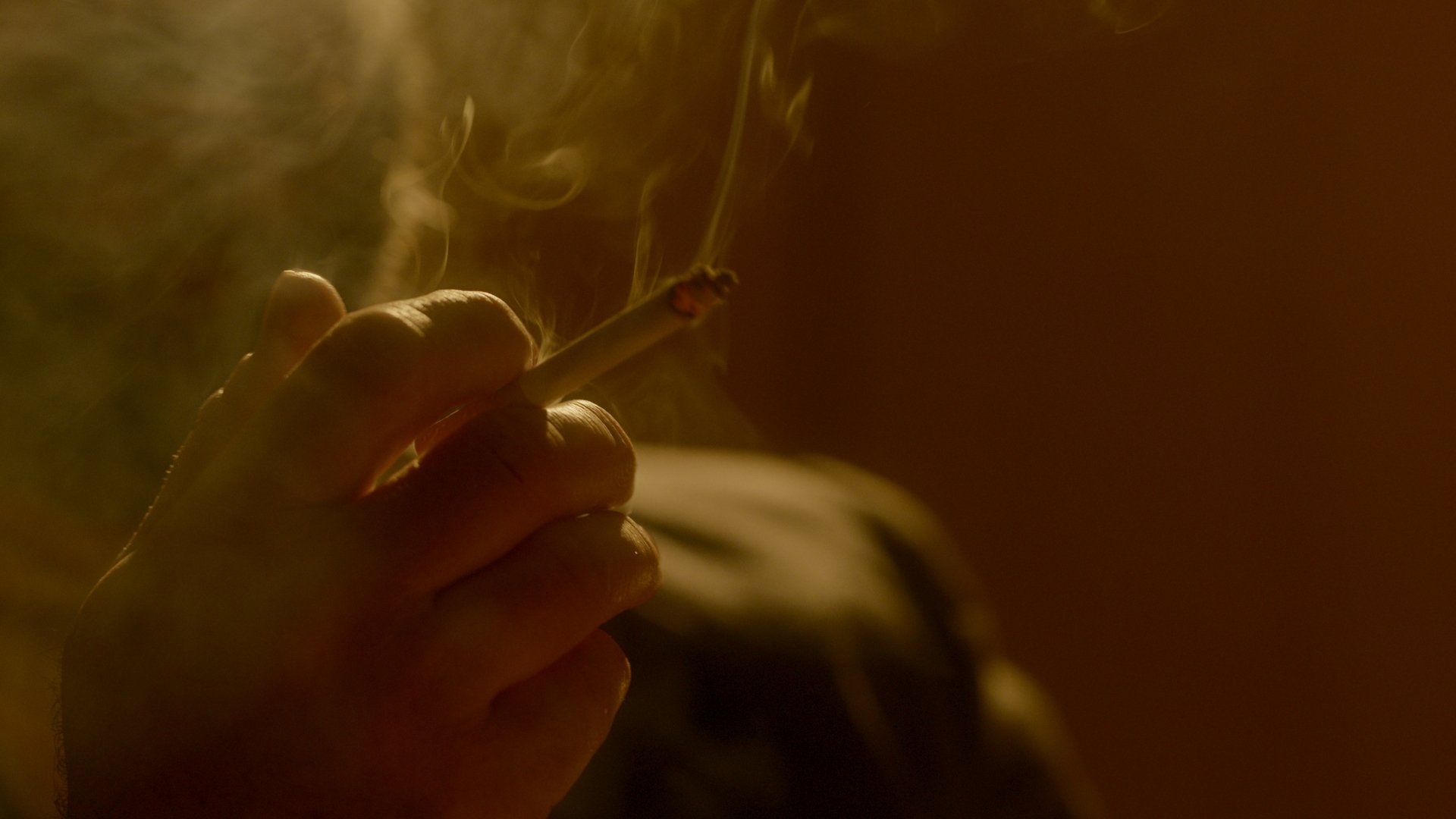 Close-up of a hand holding a lit cigarette with smoke swirling around