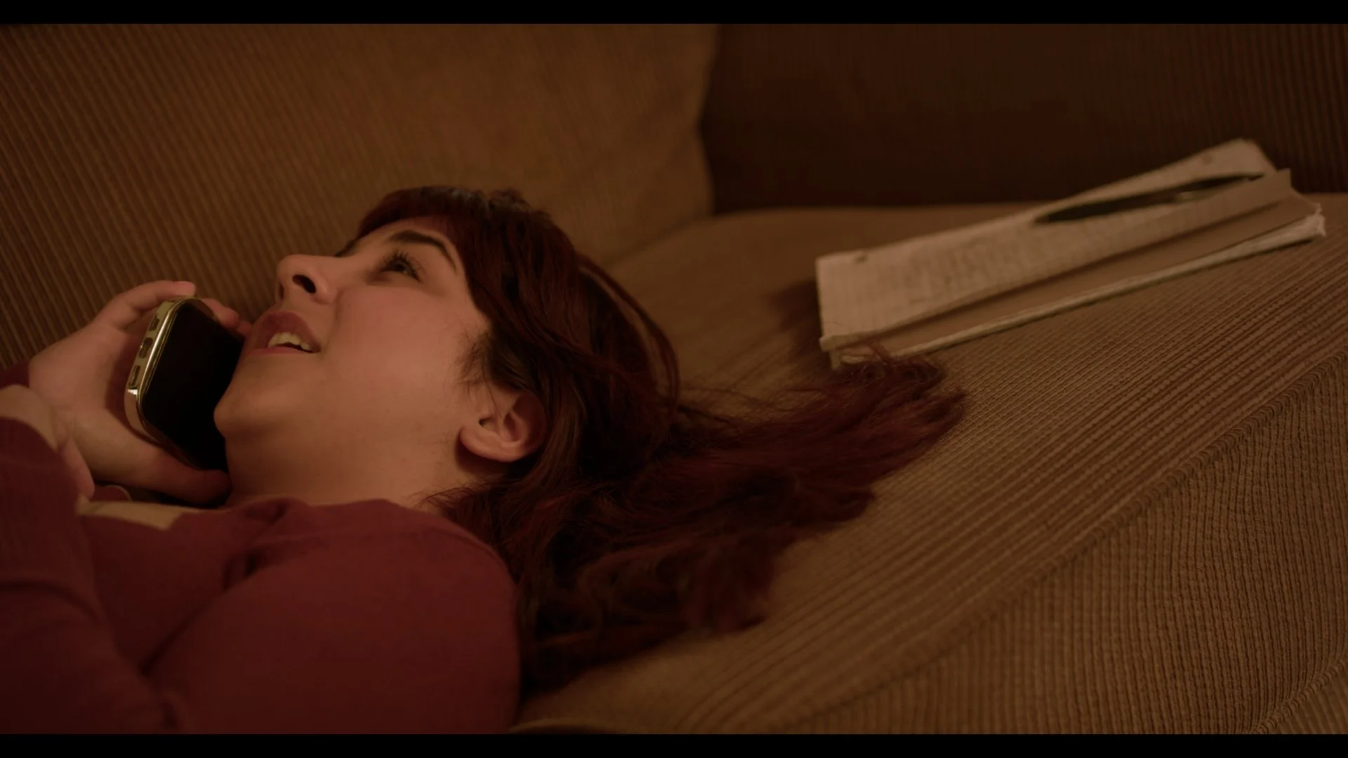 A woman with brown hair lying on a brown couch, talking on a cordless phone, with a phone book nearby.