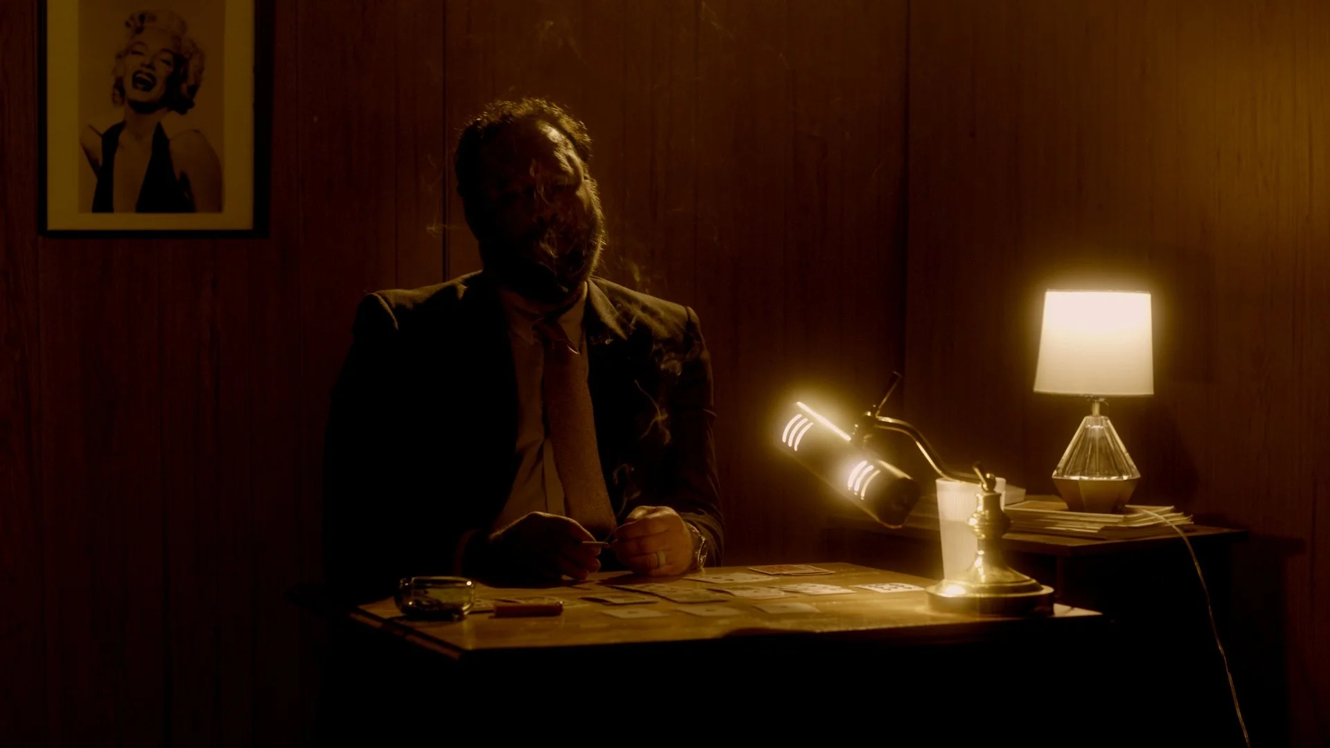 A man in a suit and tie sits at a desk in a dimly lit room, with a dark wooden wall behind him.