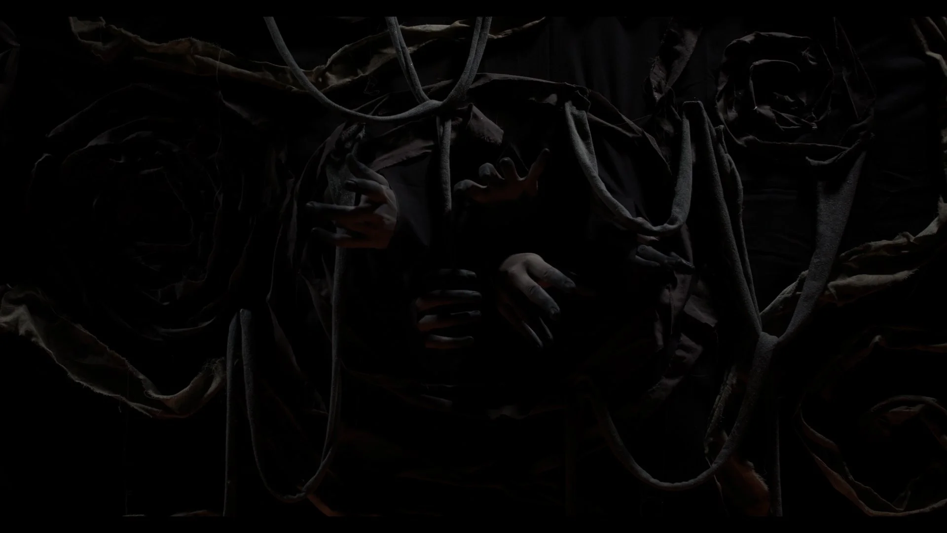 Dark image showing multiple hands reaching out from fabric or cloth surrounding a central area, creating a mysterious or eerie scene.