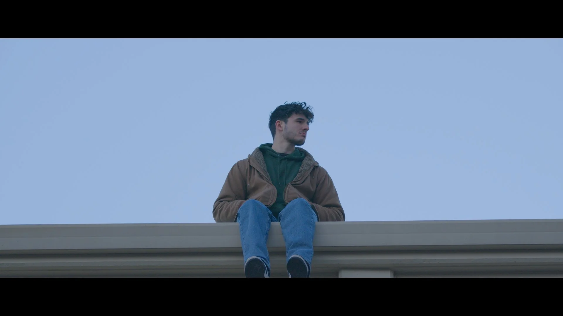 A young man sitting on a ledge against a clear blue sky, looking to the side with a thoughtful expression.