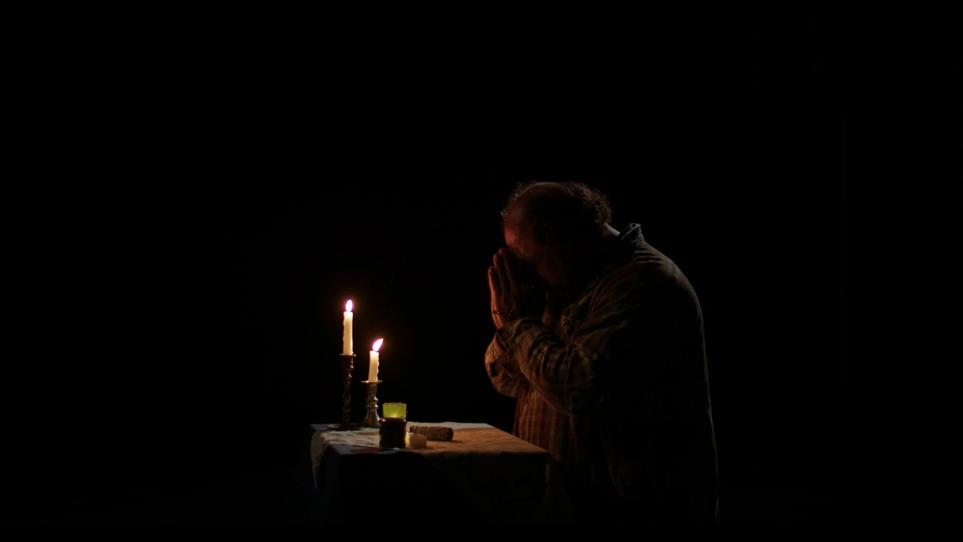 A person praying at a table with lit candles and candles on a dark background.