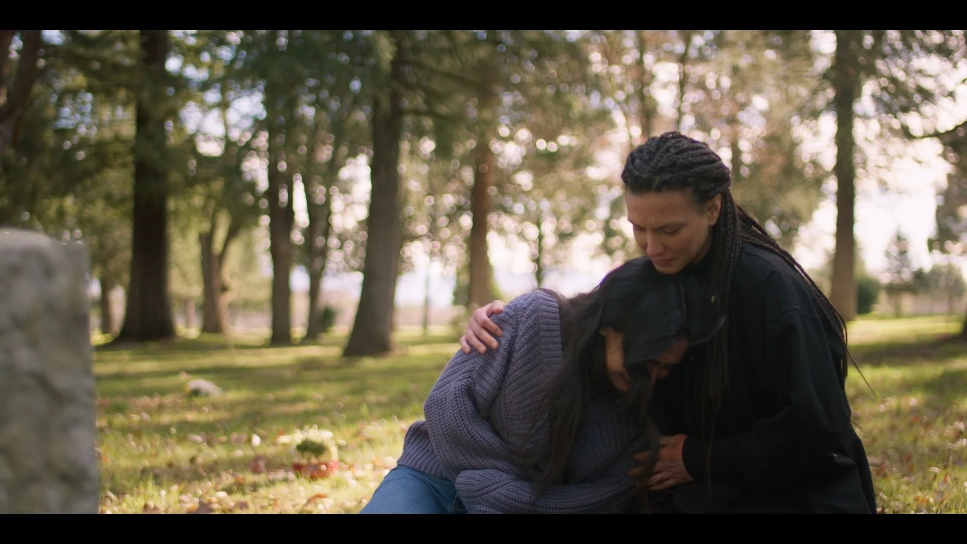 Two women comforting each other outdoors in a park during daylight, with trees in the background.
