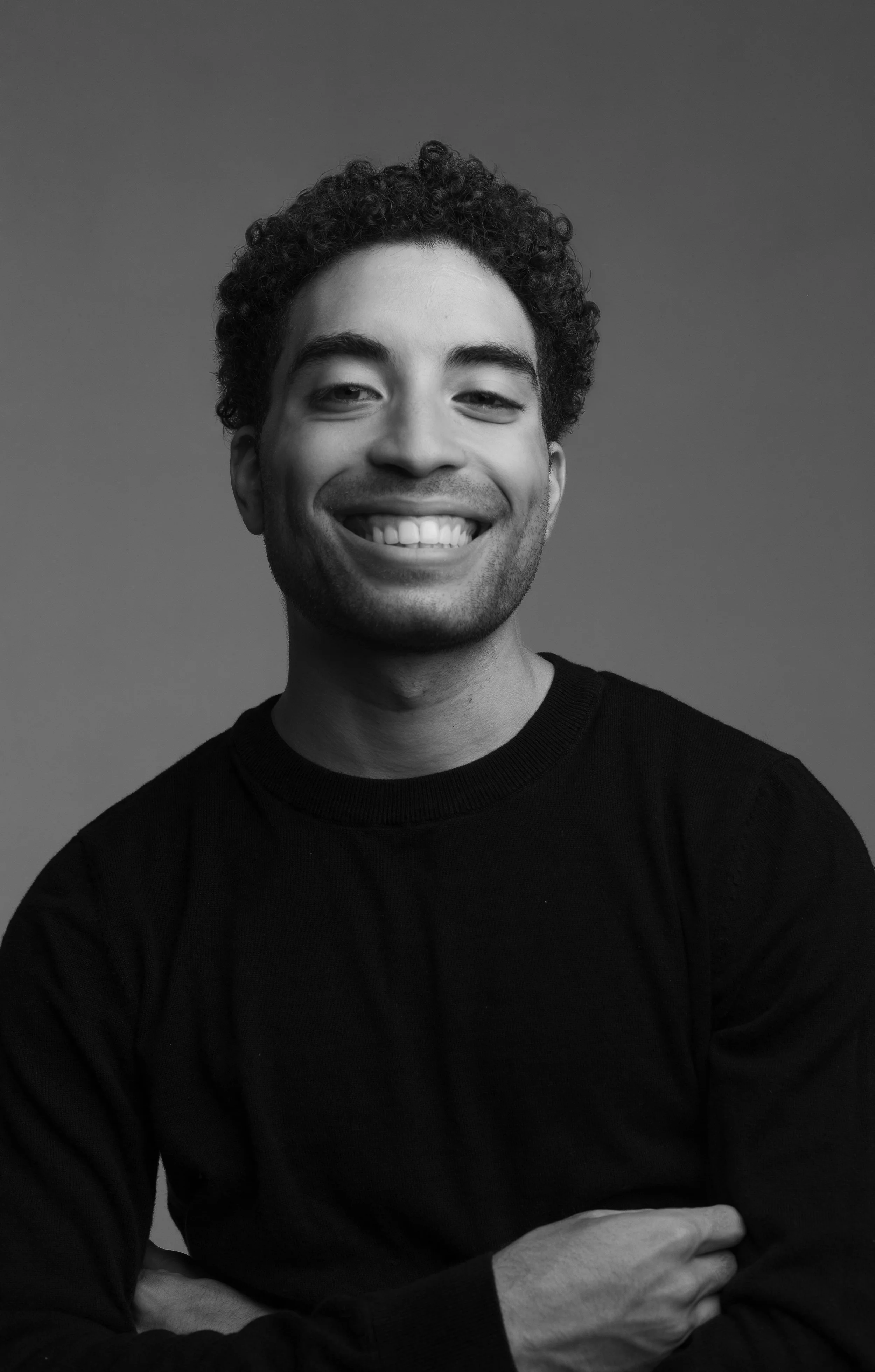 Black and white portrait of a smiling man with curly hair, wearing a black sweater, crossing his arms, against a plain background.