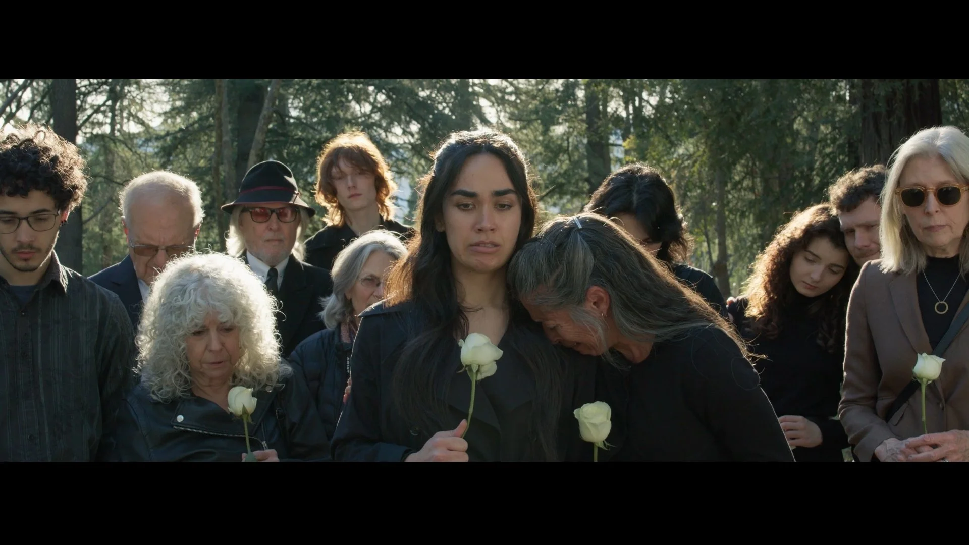 People gathered at a memorial or funeral, holding white roses, with somber expressions, standing outdoors among trees.