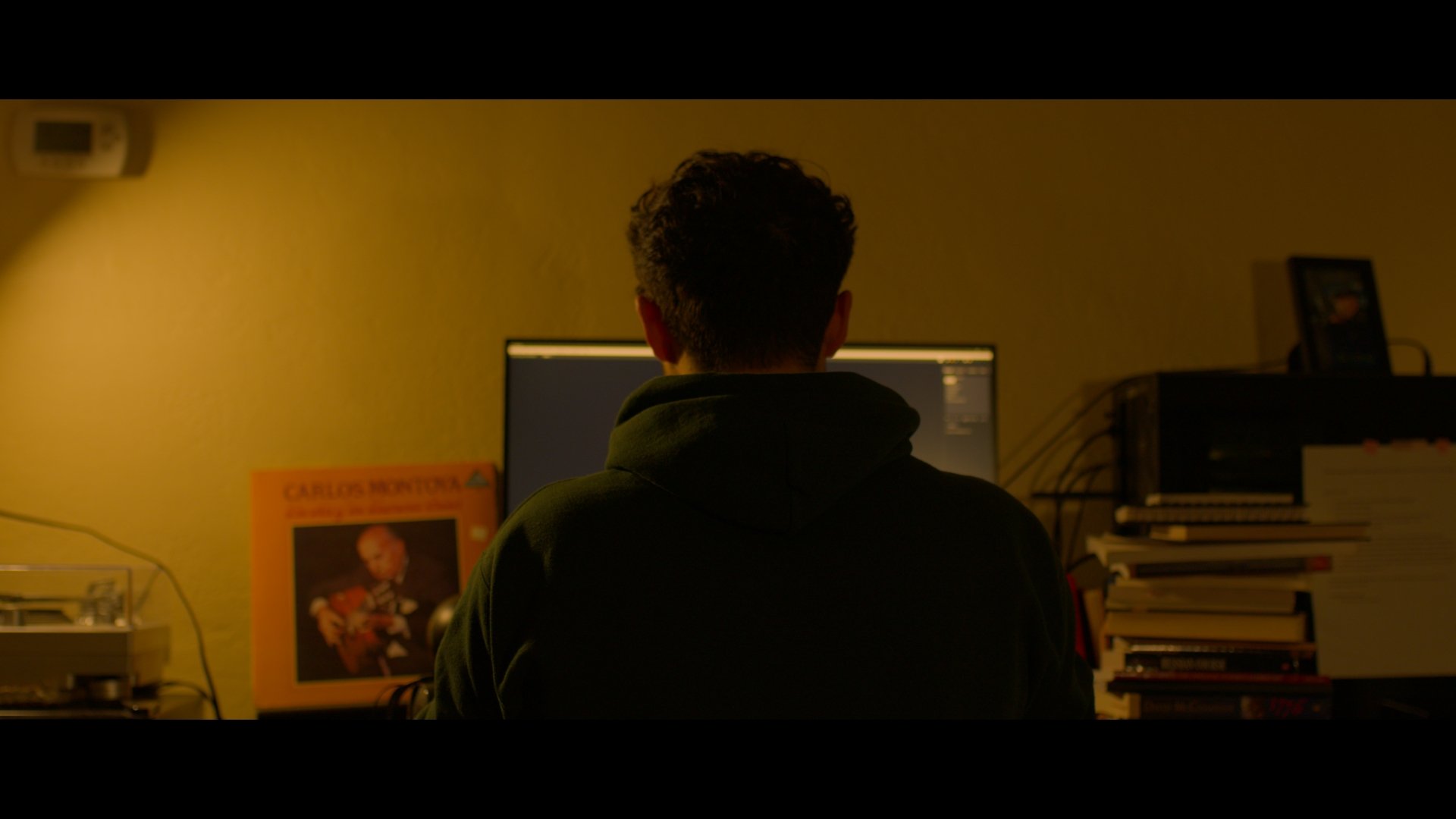 Person sitting at a desk working on a computer in a dimly lit room with books and artwork around.