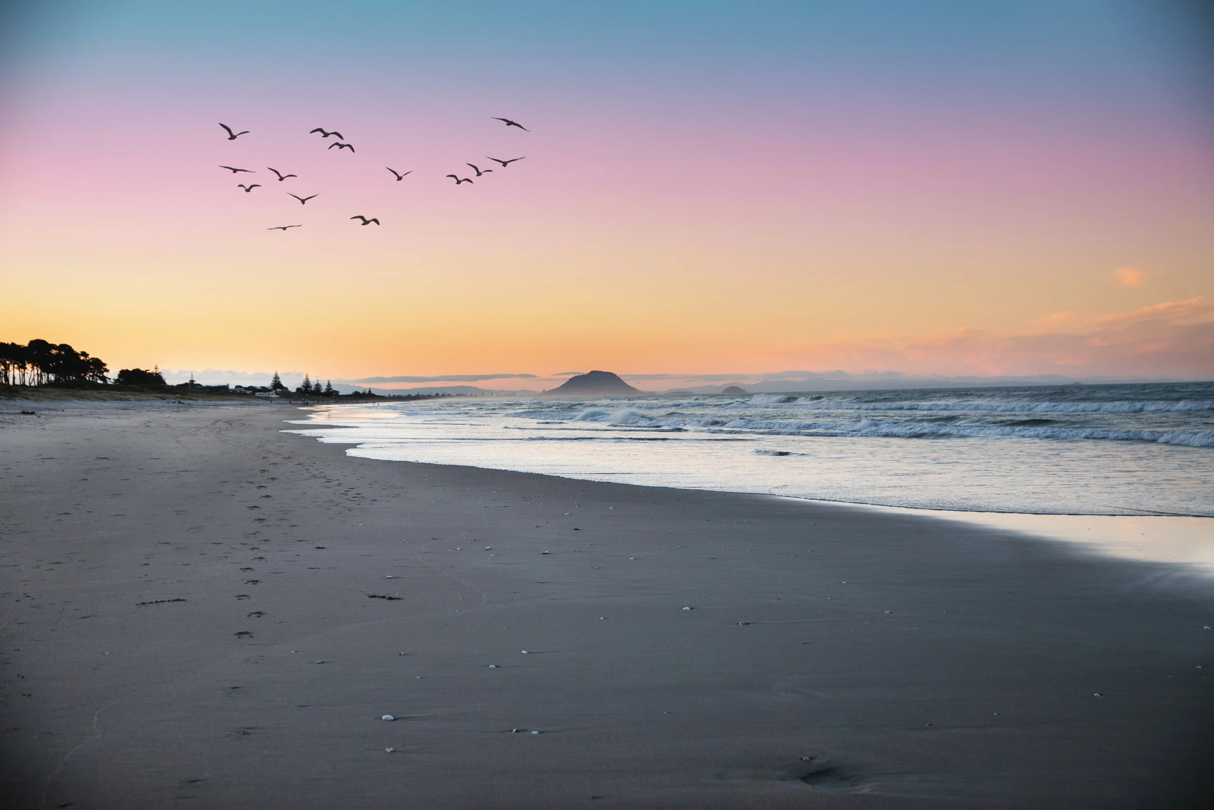 A sunset view of a Papamoa beach with birds flying in the sky and Mount Maunganui in the distance.