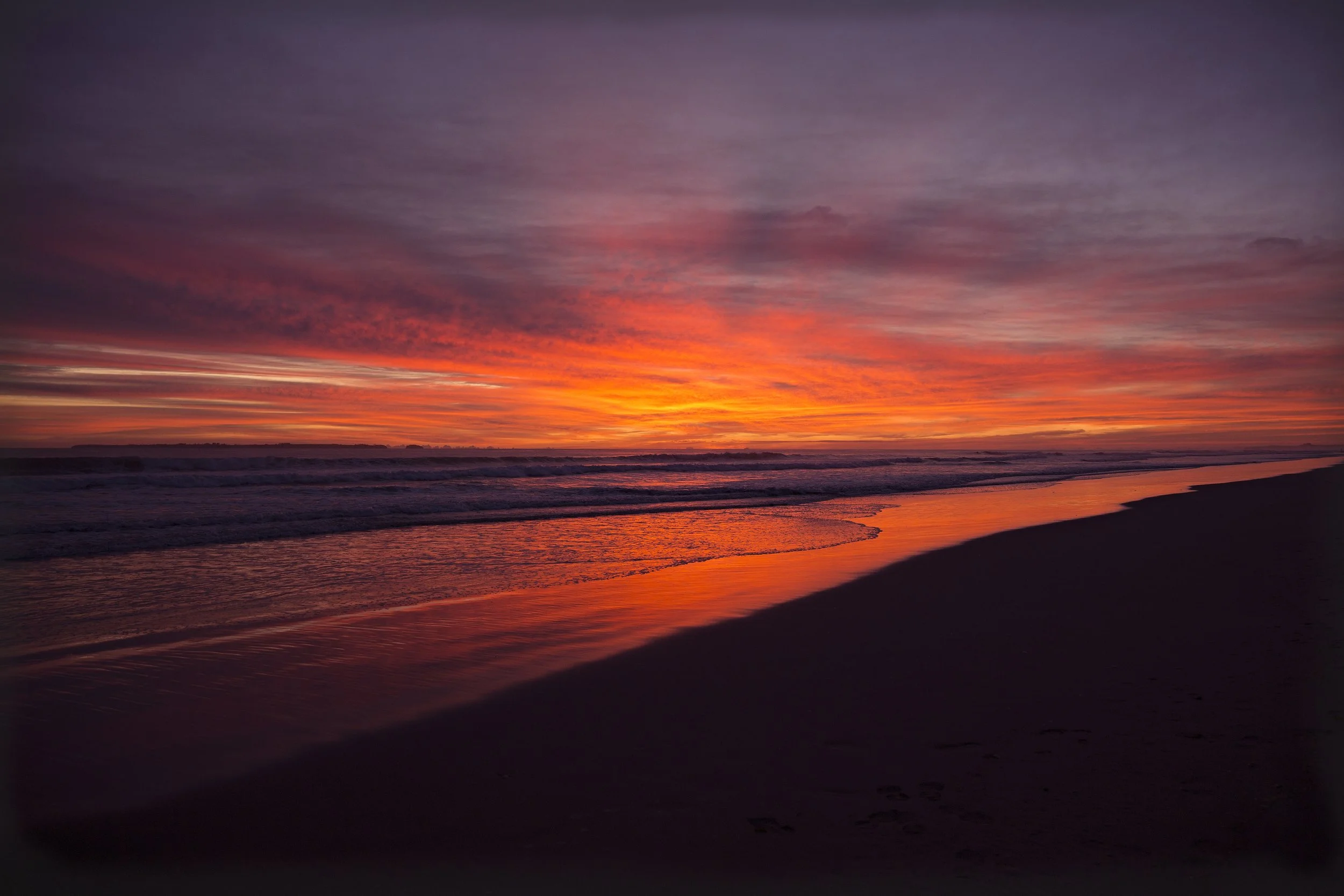 A vibrant snrise over the ocean with colorful clouds and reflections on the water, with a silhouetted shoreline in the foreground.