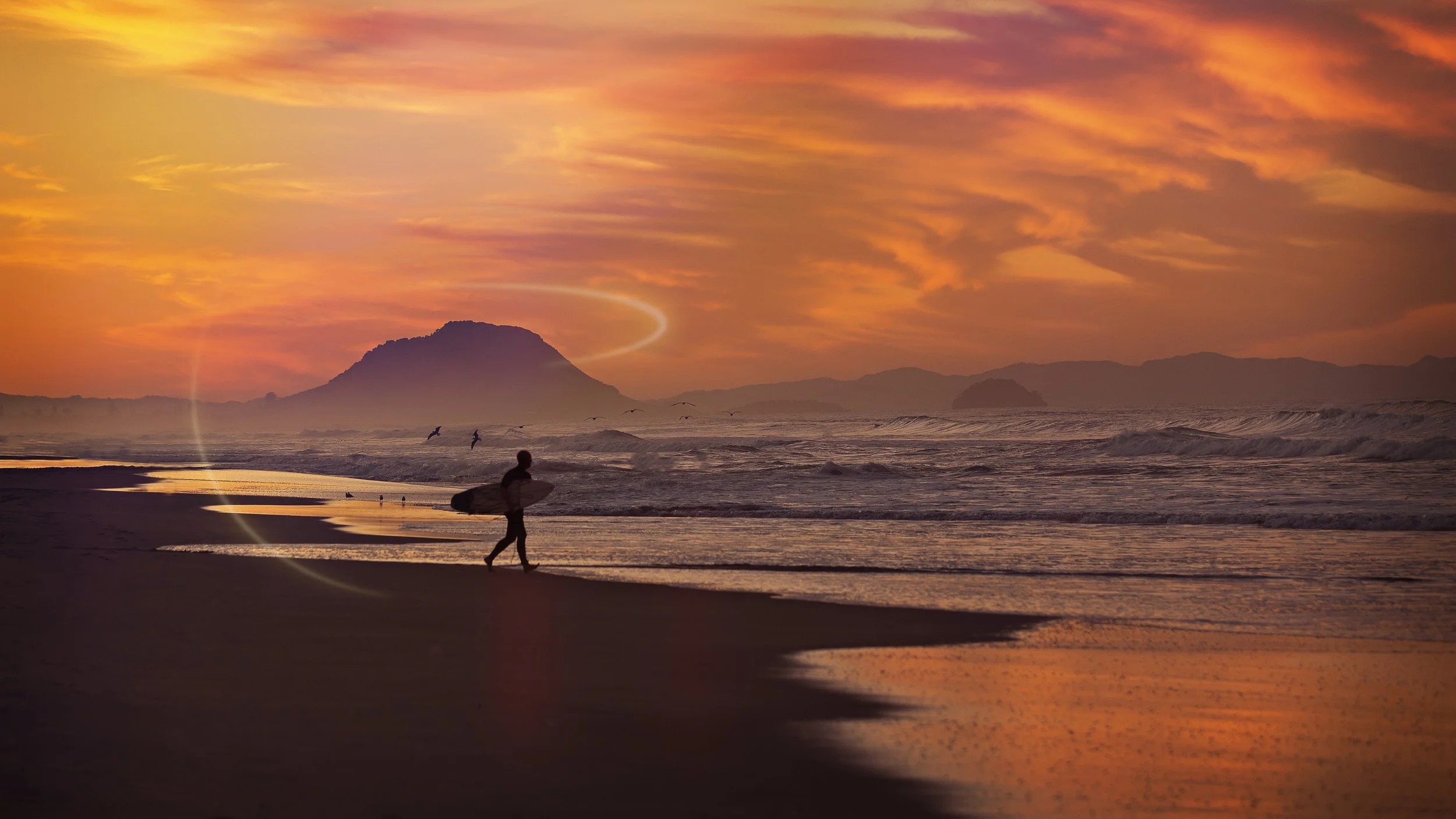 A surfer walking along the beach at sunset, with Mount Maunganui in the distance and a colorful sky above.