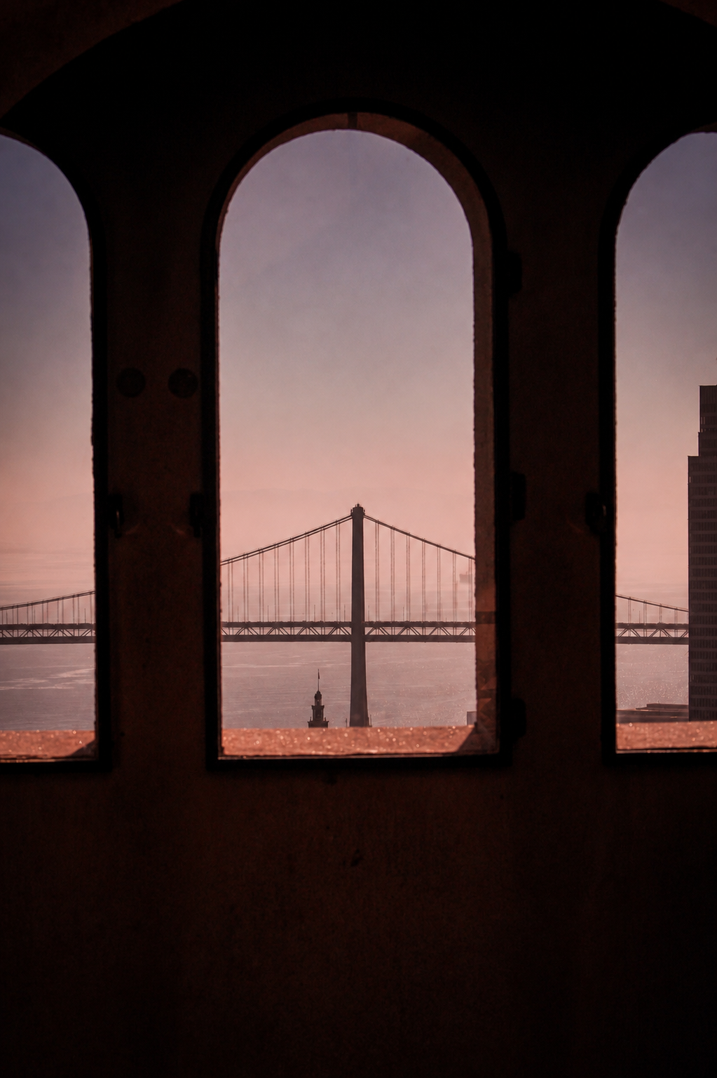 View of a suspension bridge through three arched windows with a city skyline in the background during sunset.