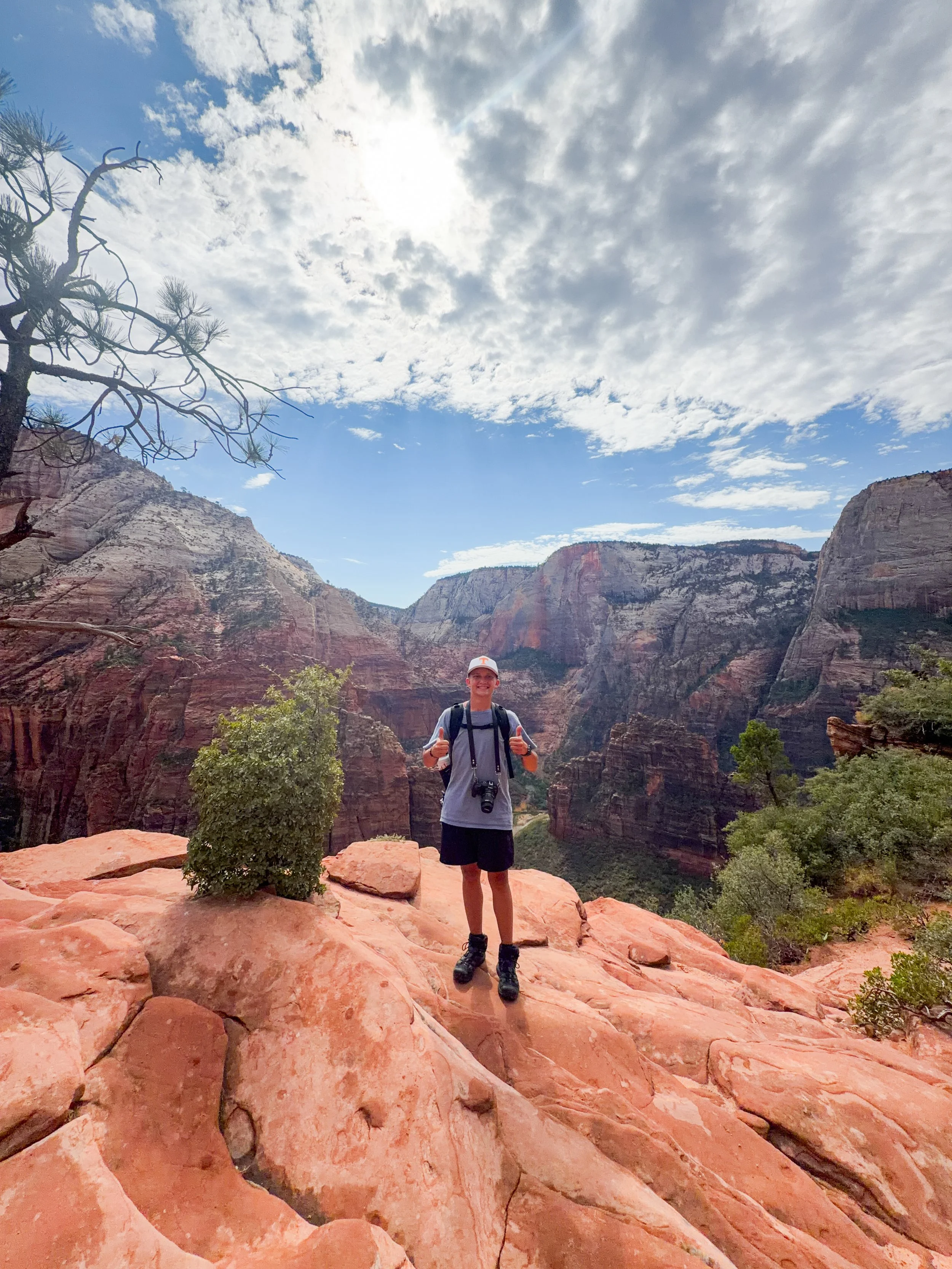 A young man with a camera around his neck and backpack gives a thumbs-up while standing on red rocks at the Grand Canyon, with canyon walls and a partly cloudy sky in the background.