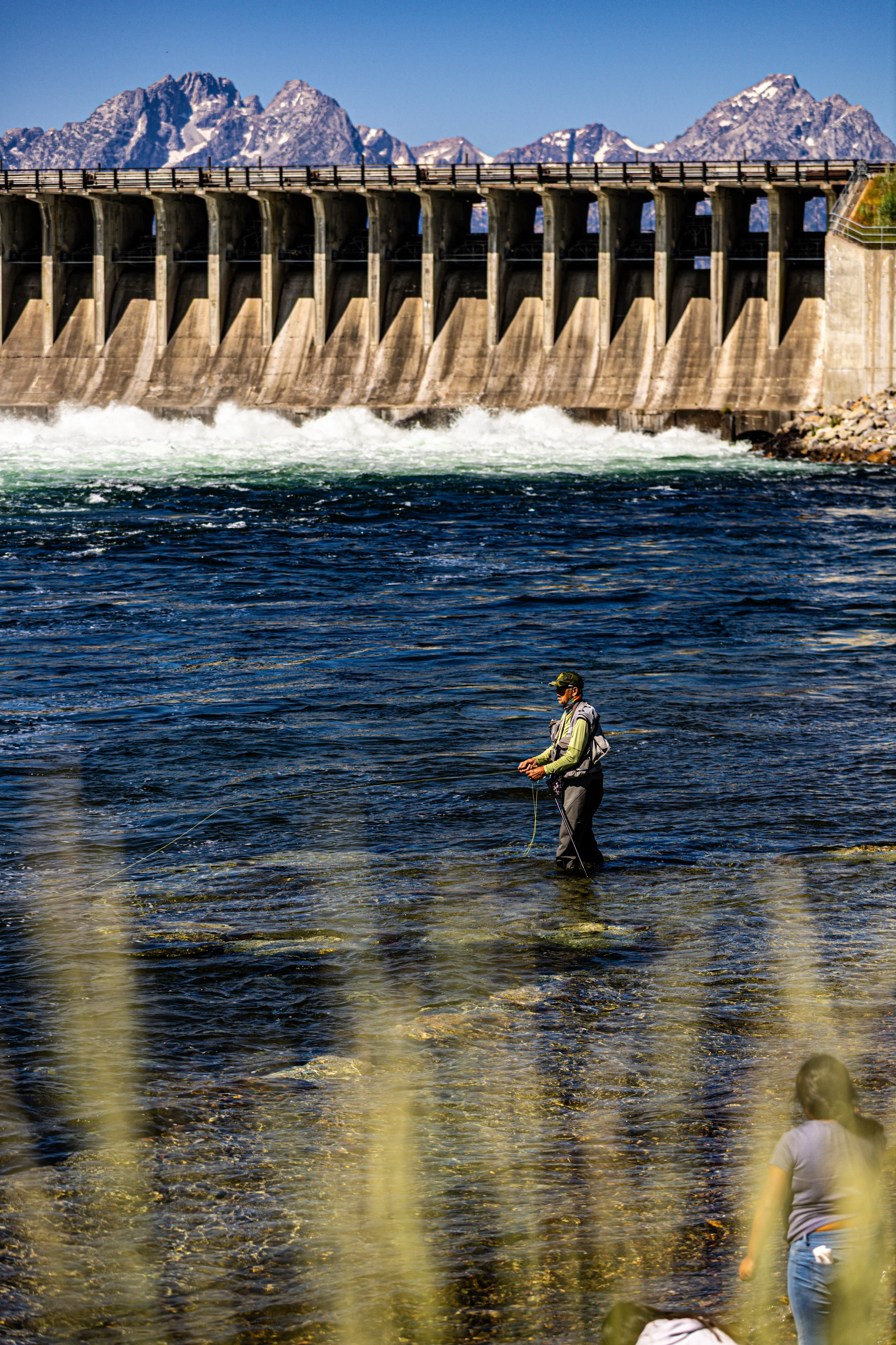 A person fly fishing in a river, with a dam and mountainous landscape in the background.