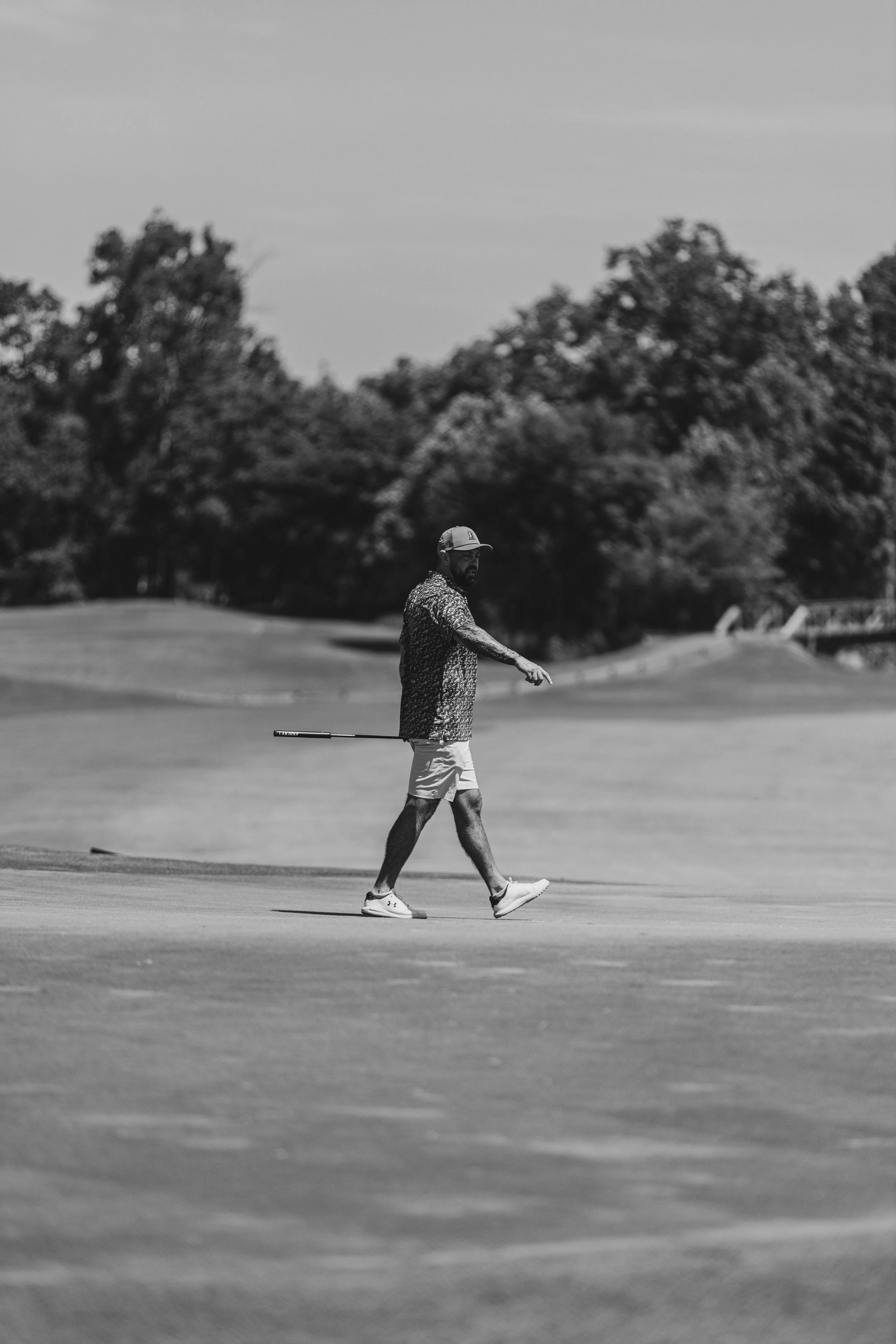 A man walking on a golf course, holding a golf club, with trees and a bridge in the background.