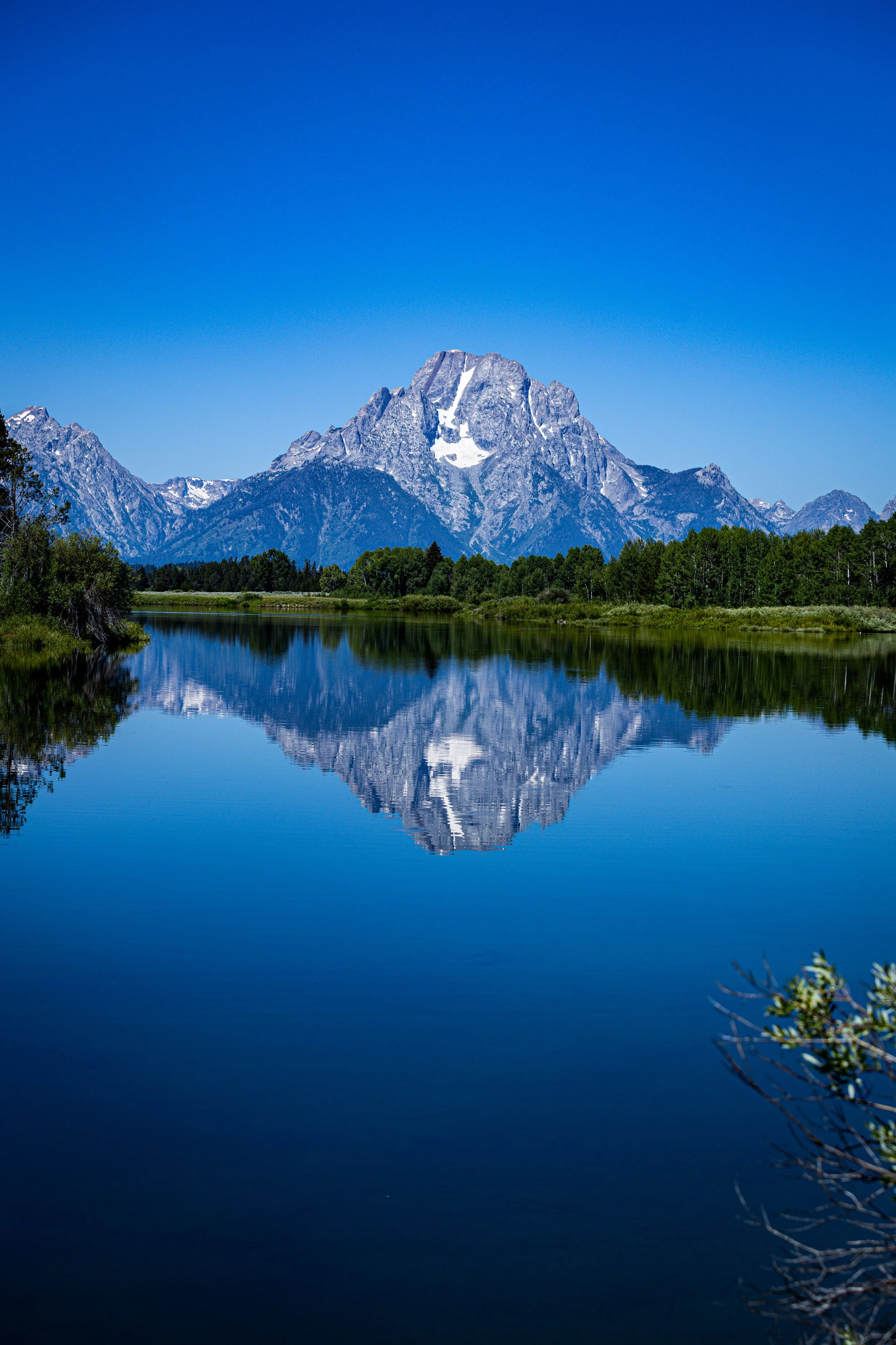A mountain with snow patches reflected in a calm lake, with green trees and clear blue sky.