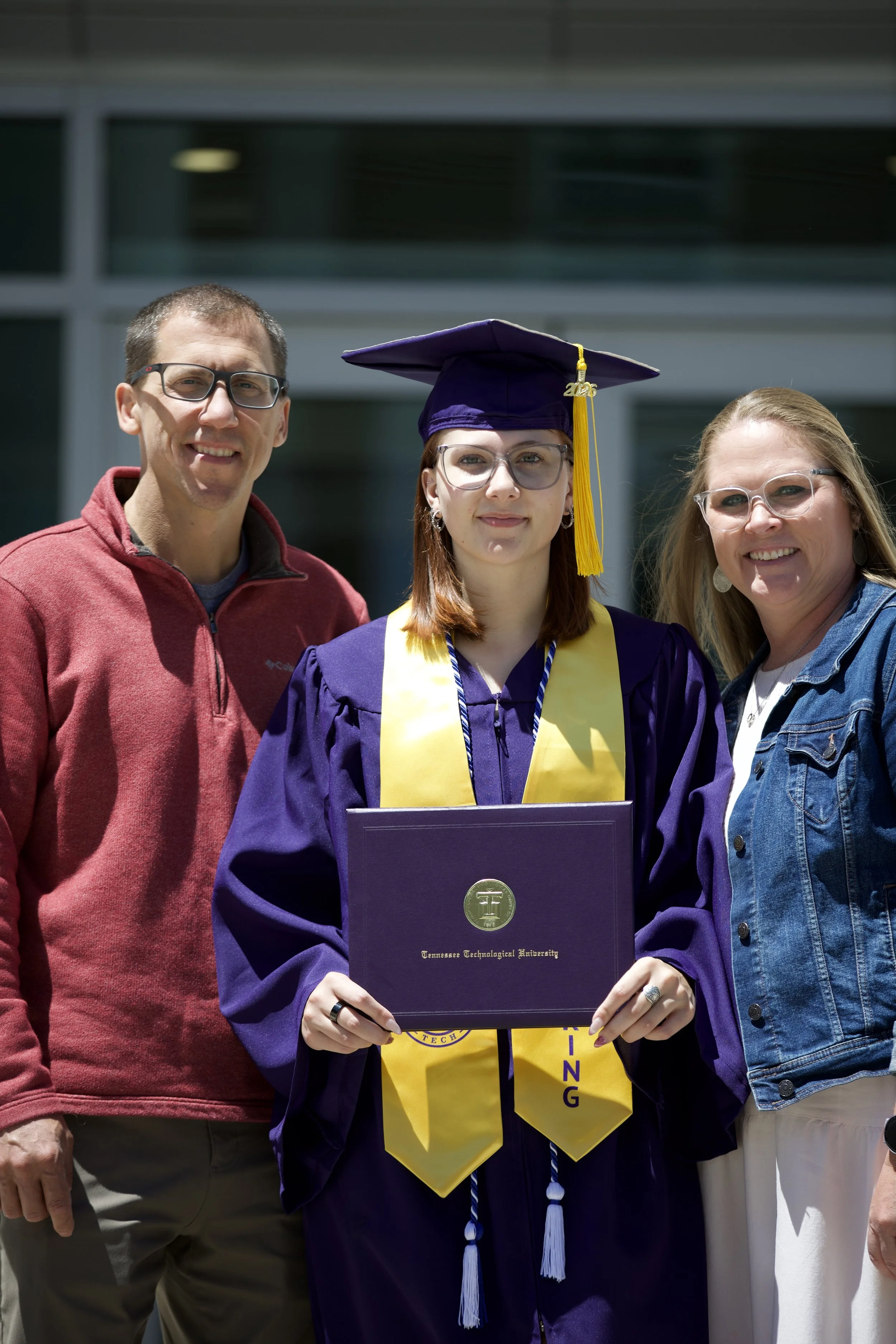 A young woman in a purple graduation cap and gown holding a diploma from Tennessee Technological University, standing outdoors with two adults on either side.
