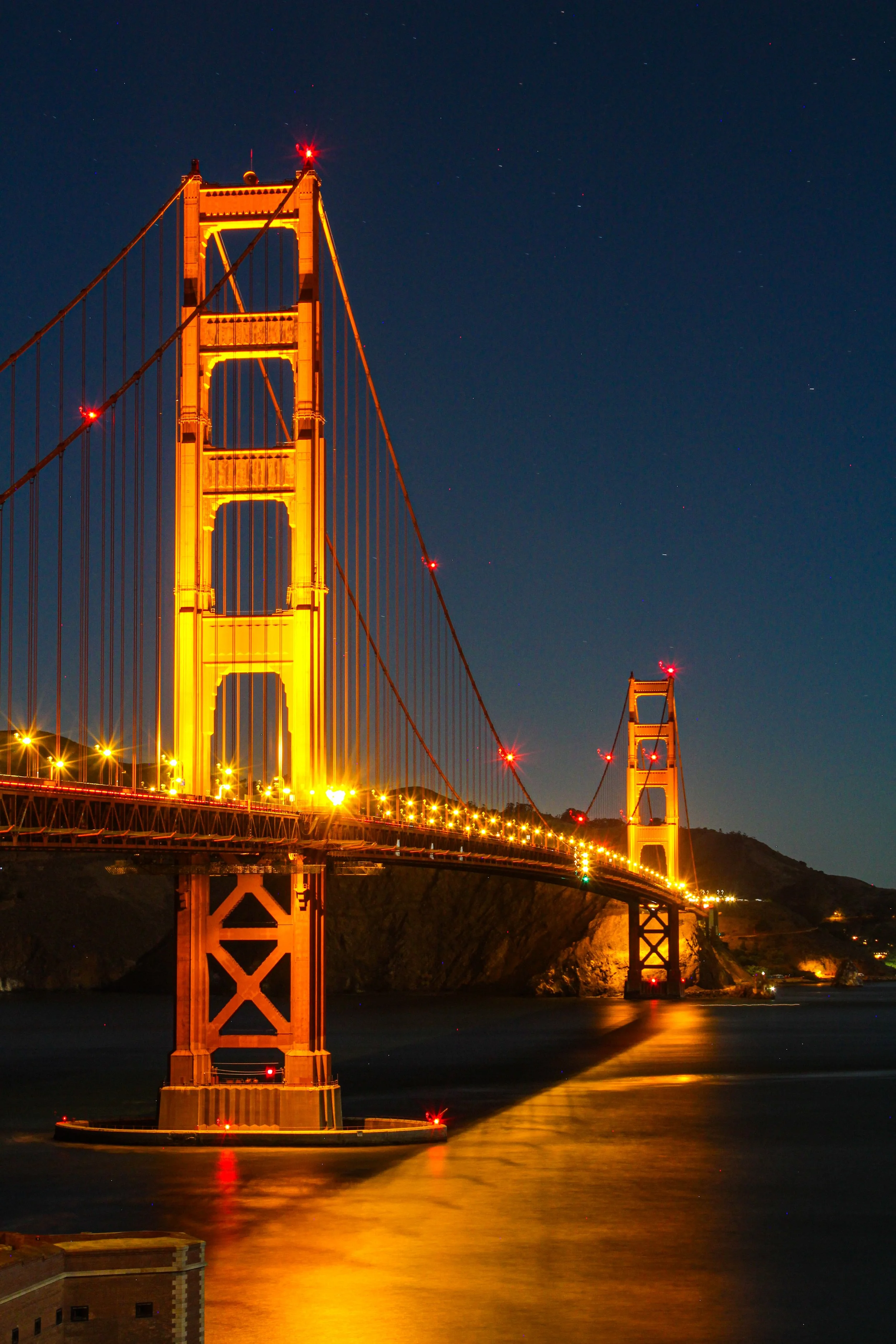 The Golden Gate Bridge at night, illuminated in orange, with city lights reflecting on the water below and stars visible in the dark sky.