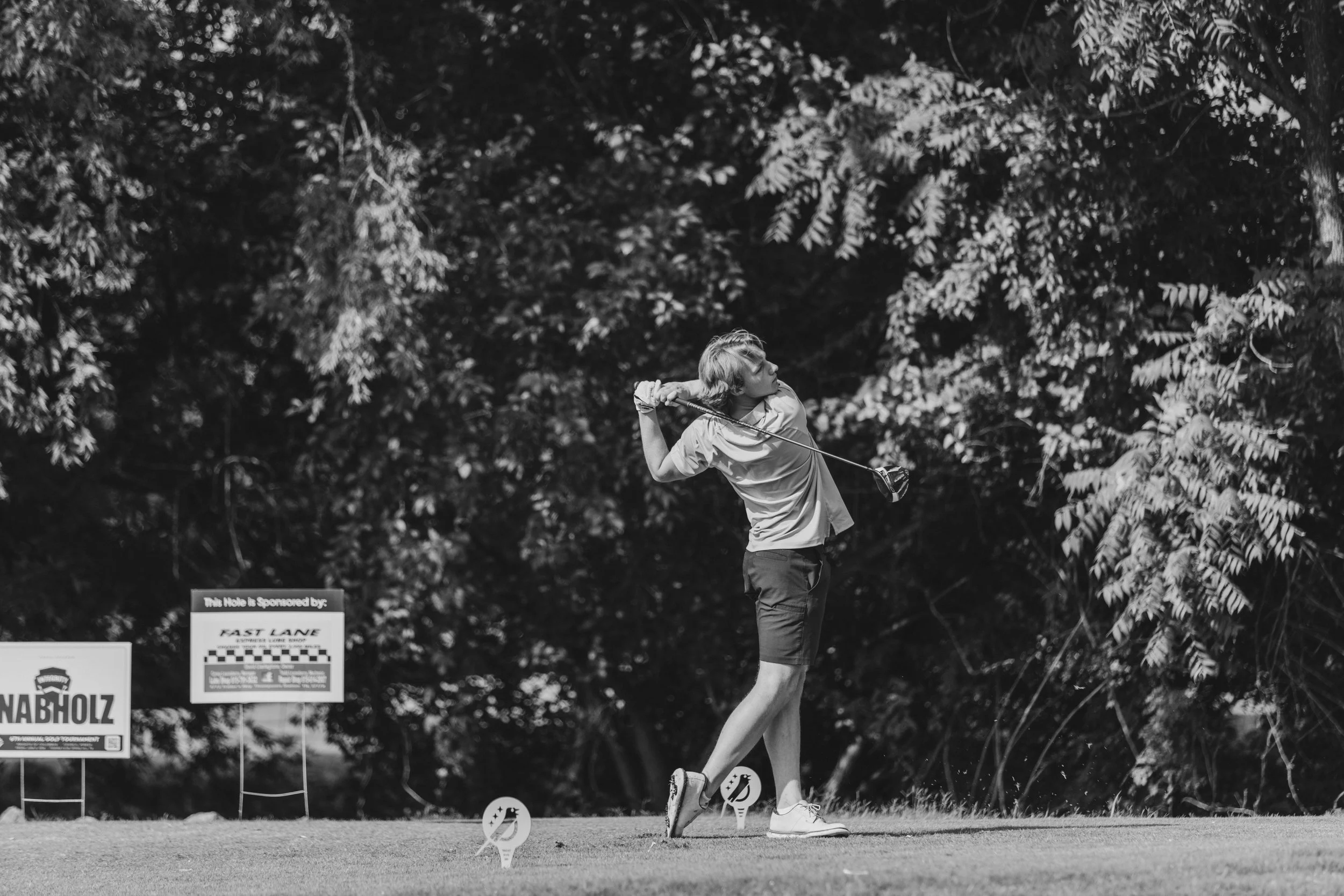 A young man in a golf course, swinging a golf club, with trees in the background and signs on the ground.