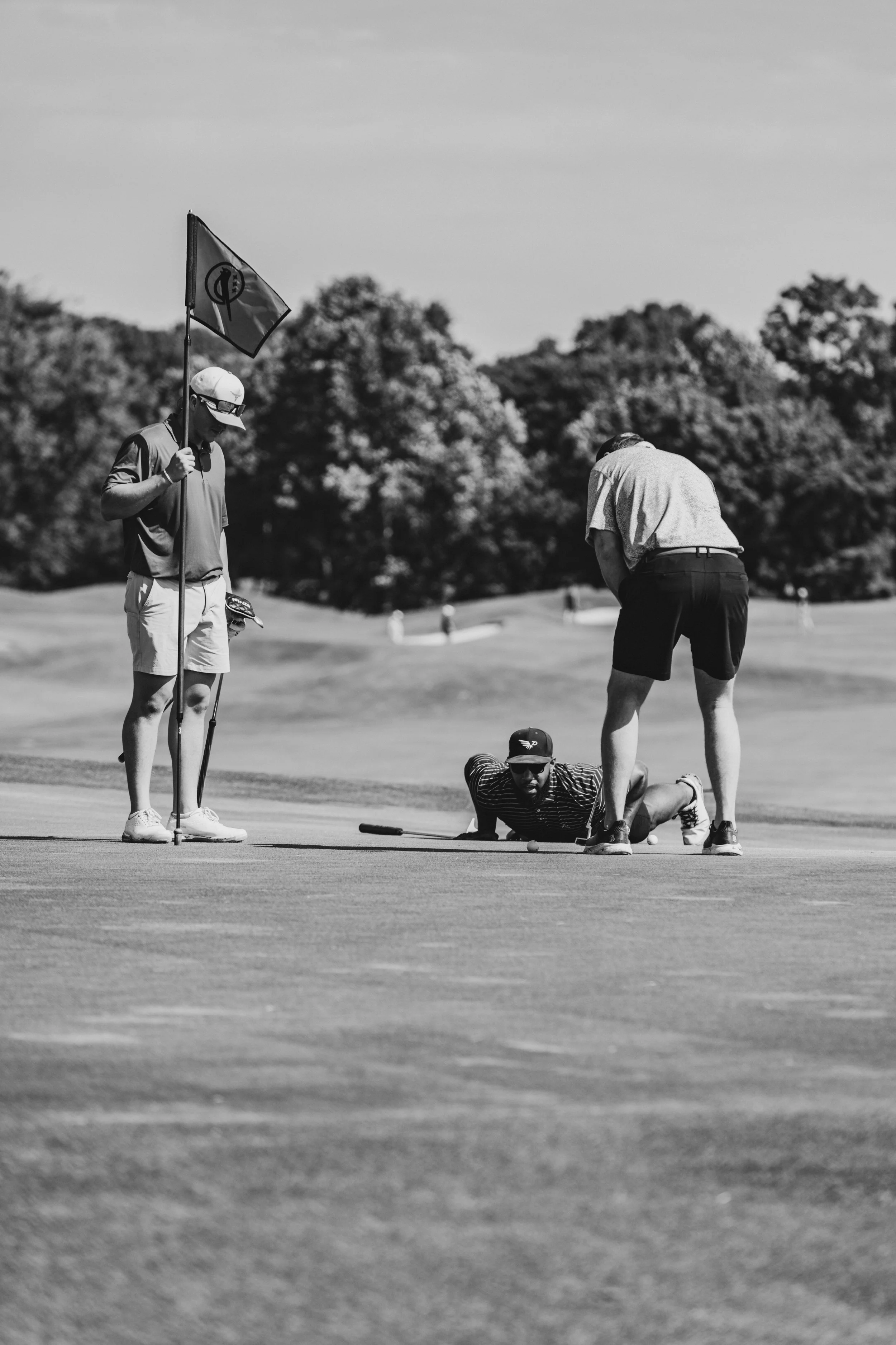 A golf caddy holding a flag near a golfer and a person lying on the ground on a golf course with trees in the background.