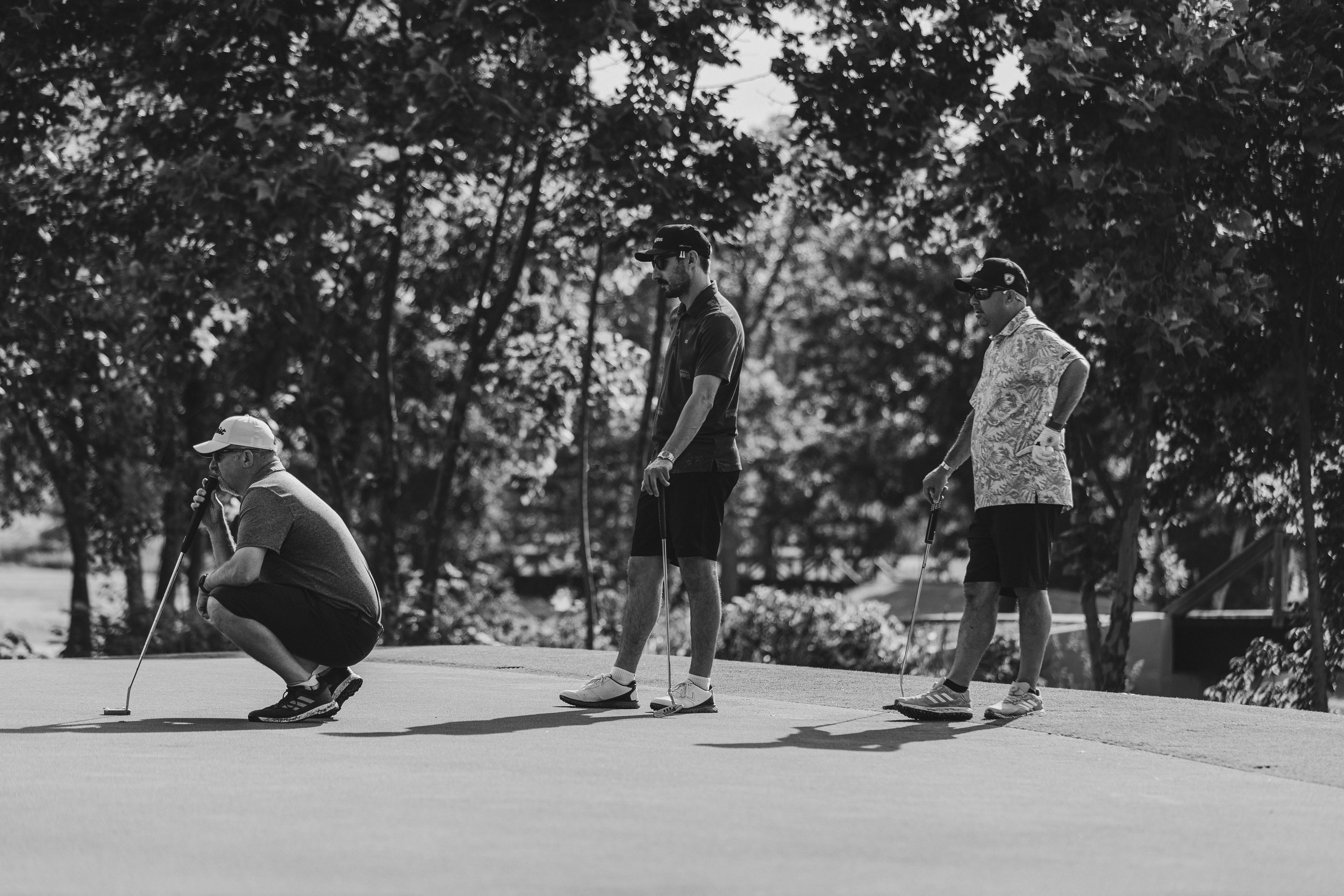 Four men playing golf on a course, with trees in the background; one man is kneeling, three others are standing, some wearing caps and sunglasses, all holding golf clubs.