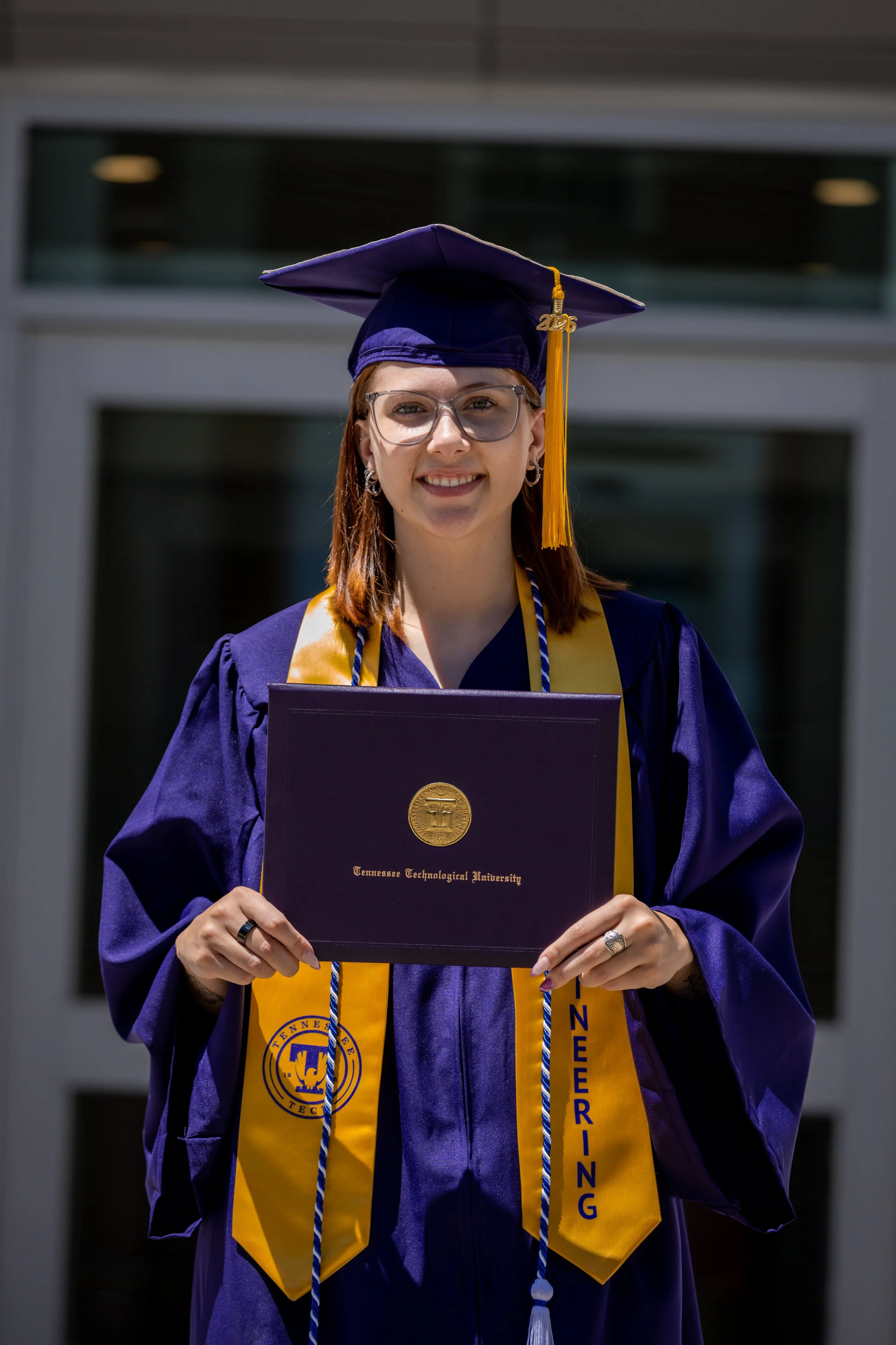 A woman wearing purple graduation gown and cap, holding a diploma from Tennessee Technological University, standing outdoors in front of a building.