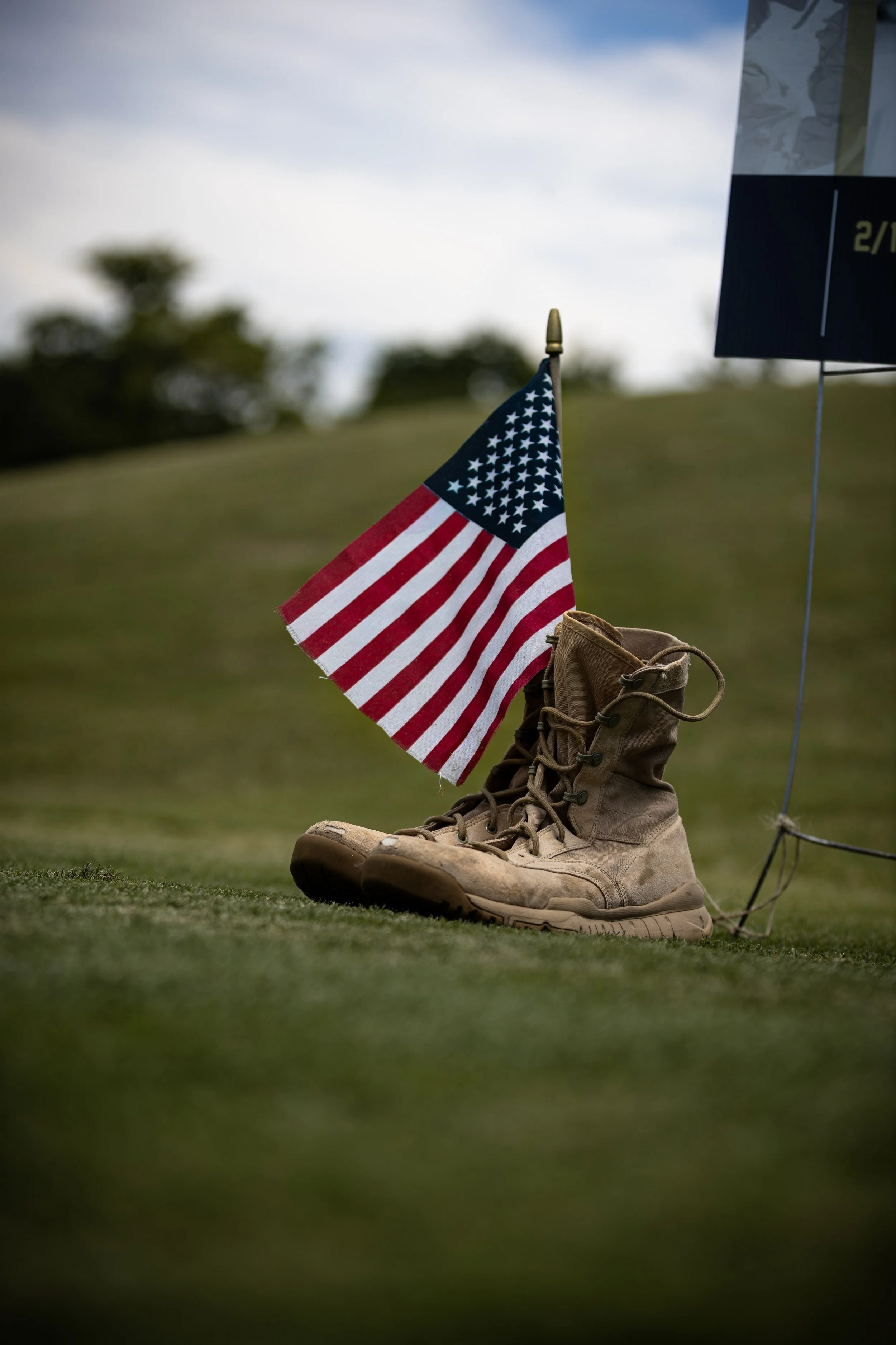 Military boots on grass with an American flag and a nearby sign or display in the background under cloudy sky.