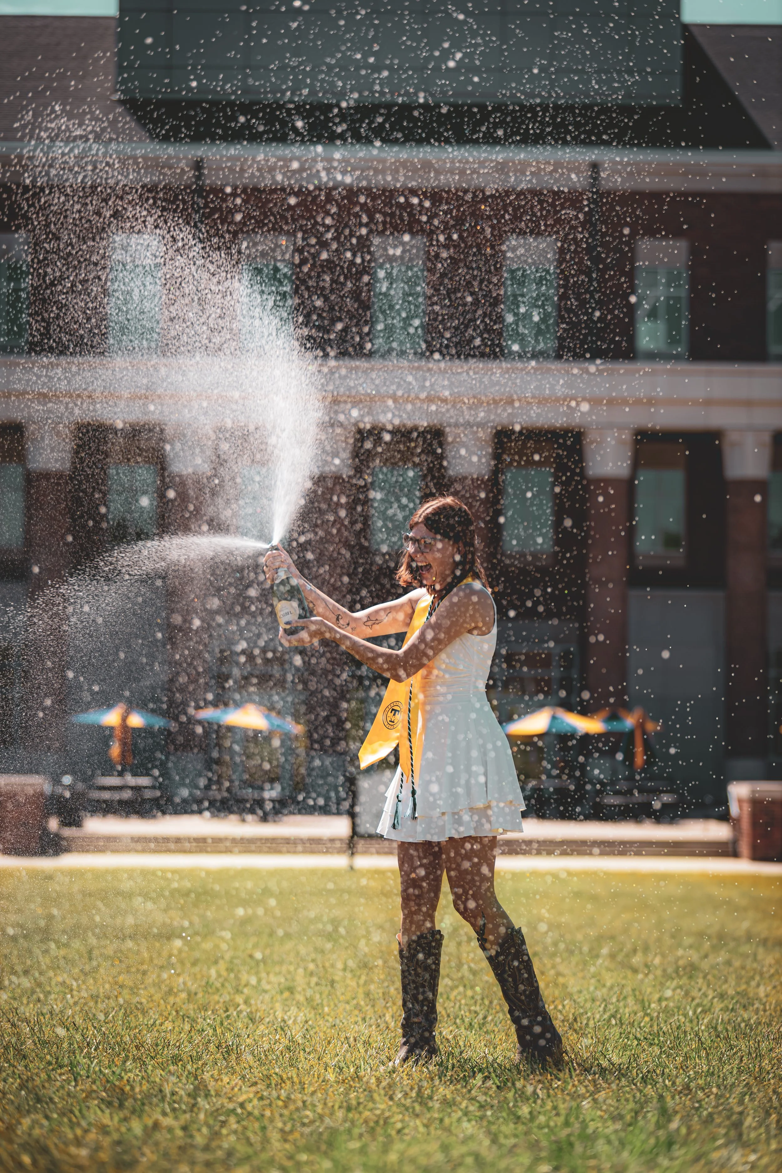 A woman wearing a white dress and cowboy boots celebrating outdoors, holding a champagne bottle and spraying champagne, with a school or office building in the background and yellow umbrellas on the grass.
