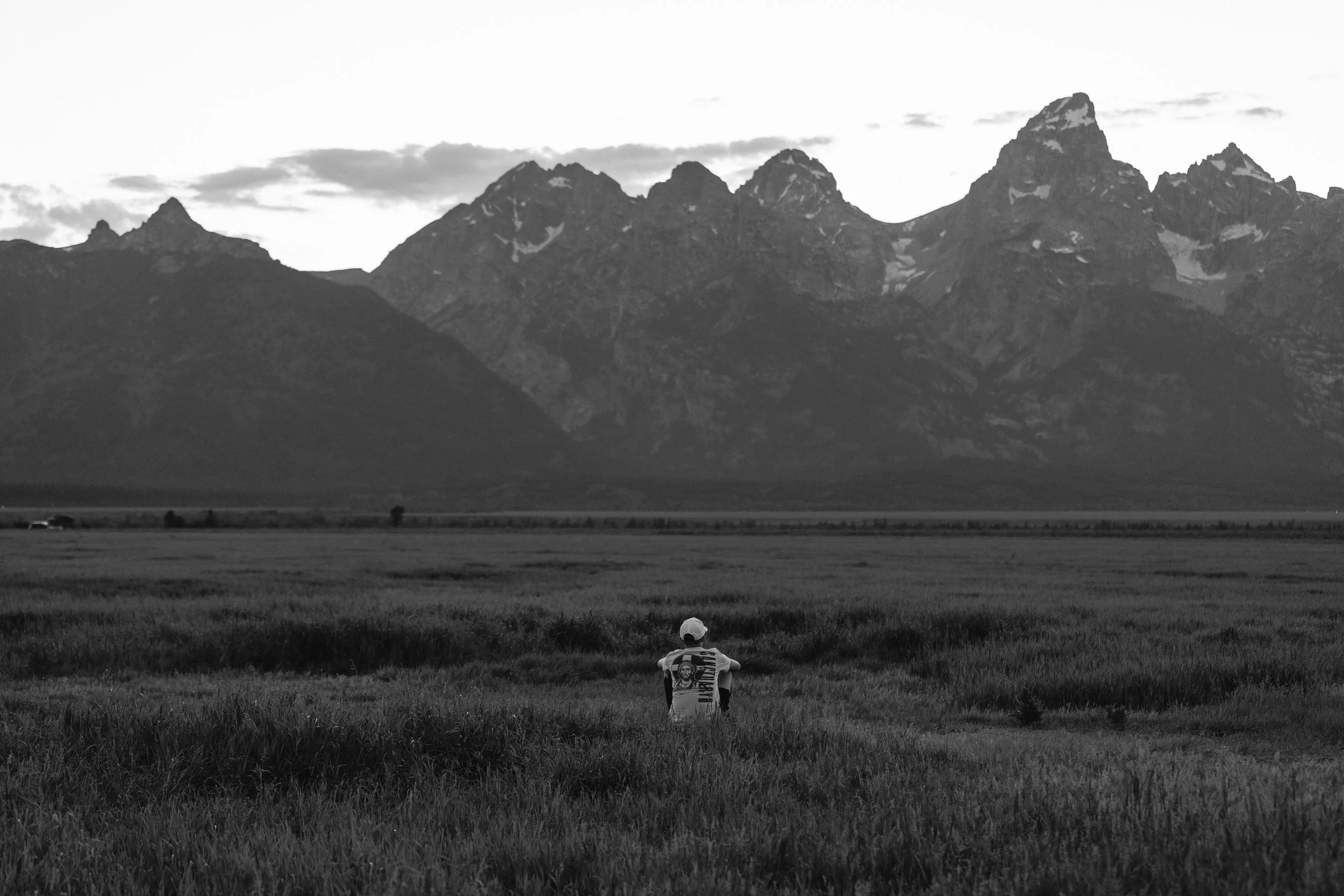 A person wearing a white hat and striped shirt sitting on a grassy plain, looking at a mountain range in the distance under a cloudy sky.