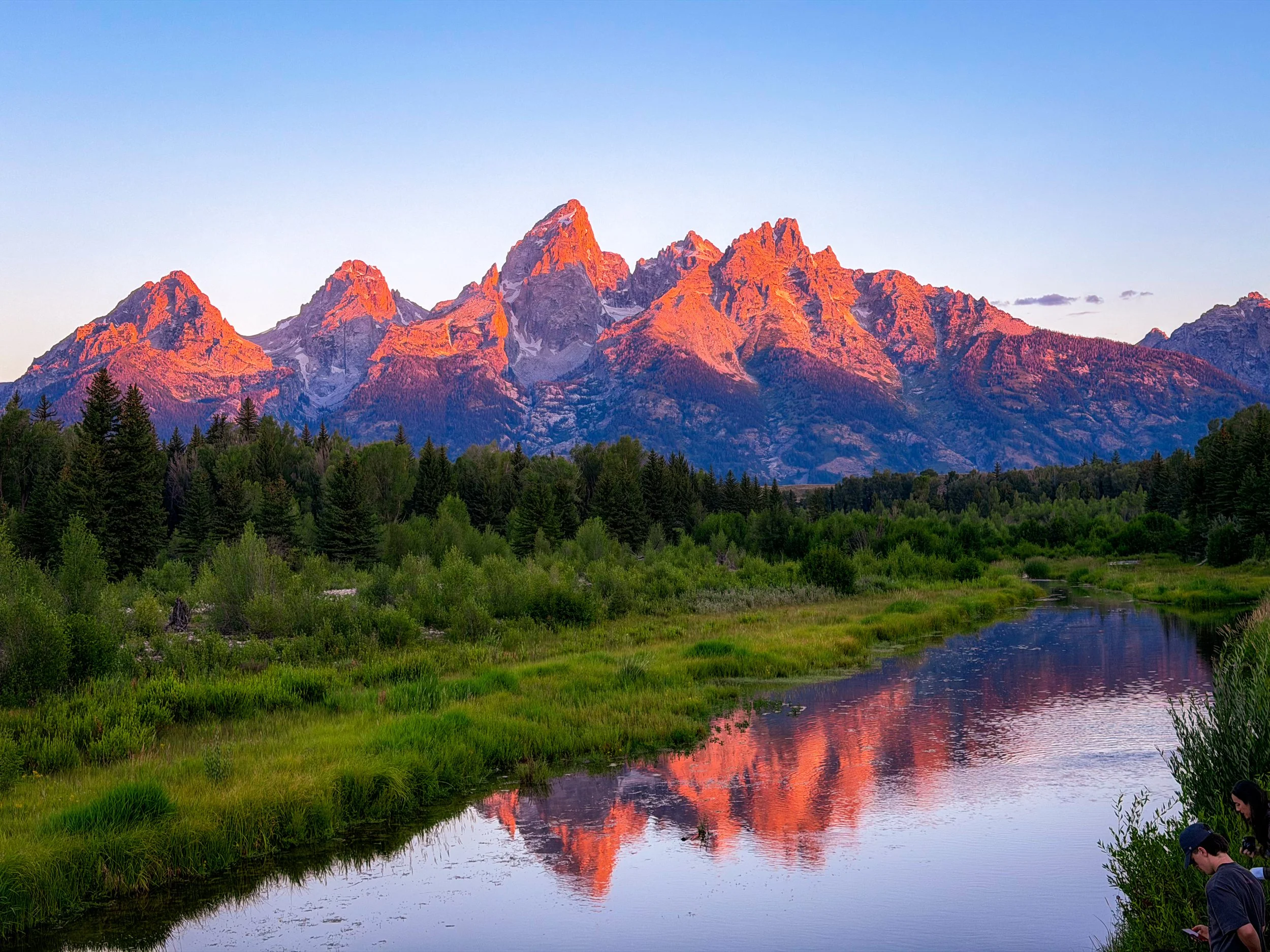 Sunset over jagged mountain peaks reflected in a calm river, with green trees in the foreground and people fishing at the riverbank.
