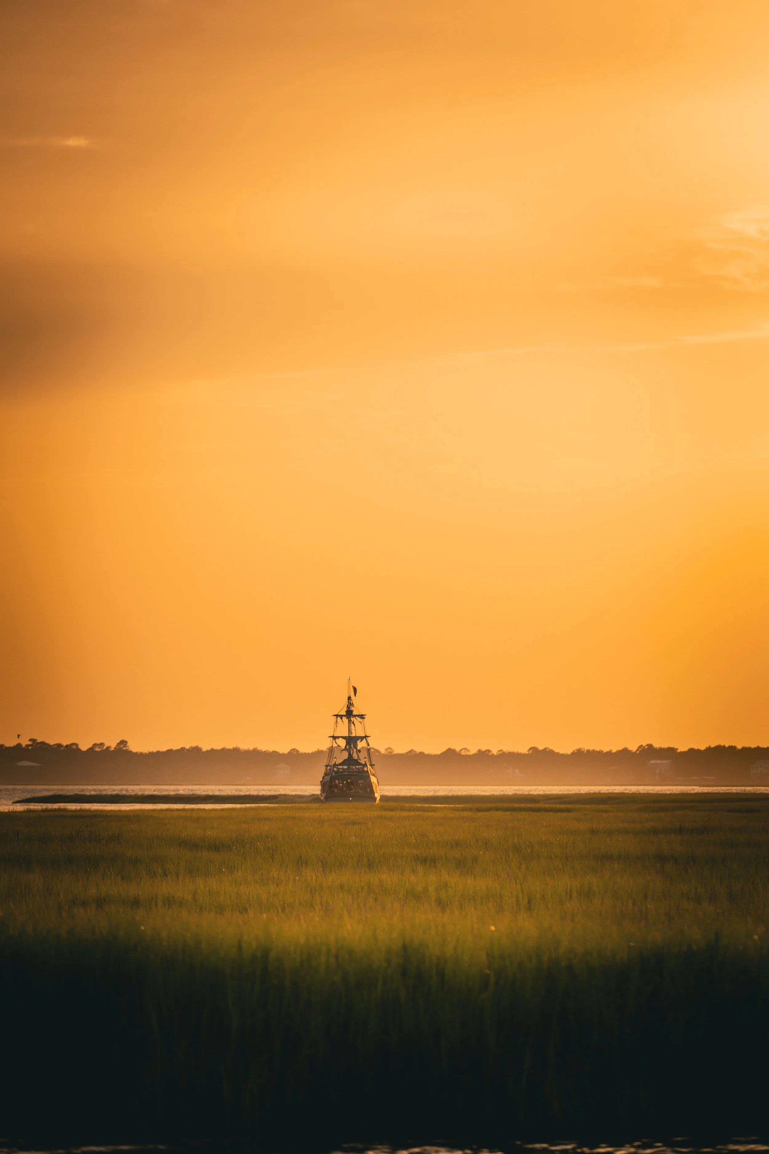 A yacht sailing on a calm body of water at sunset with a vibrant orange sky, dark silhouettes of trees in the background, and green grass in the foreground.