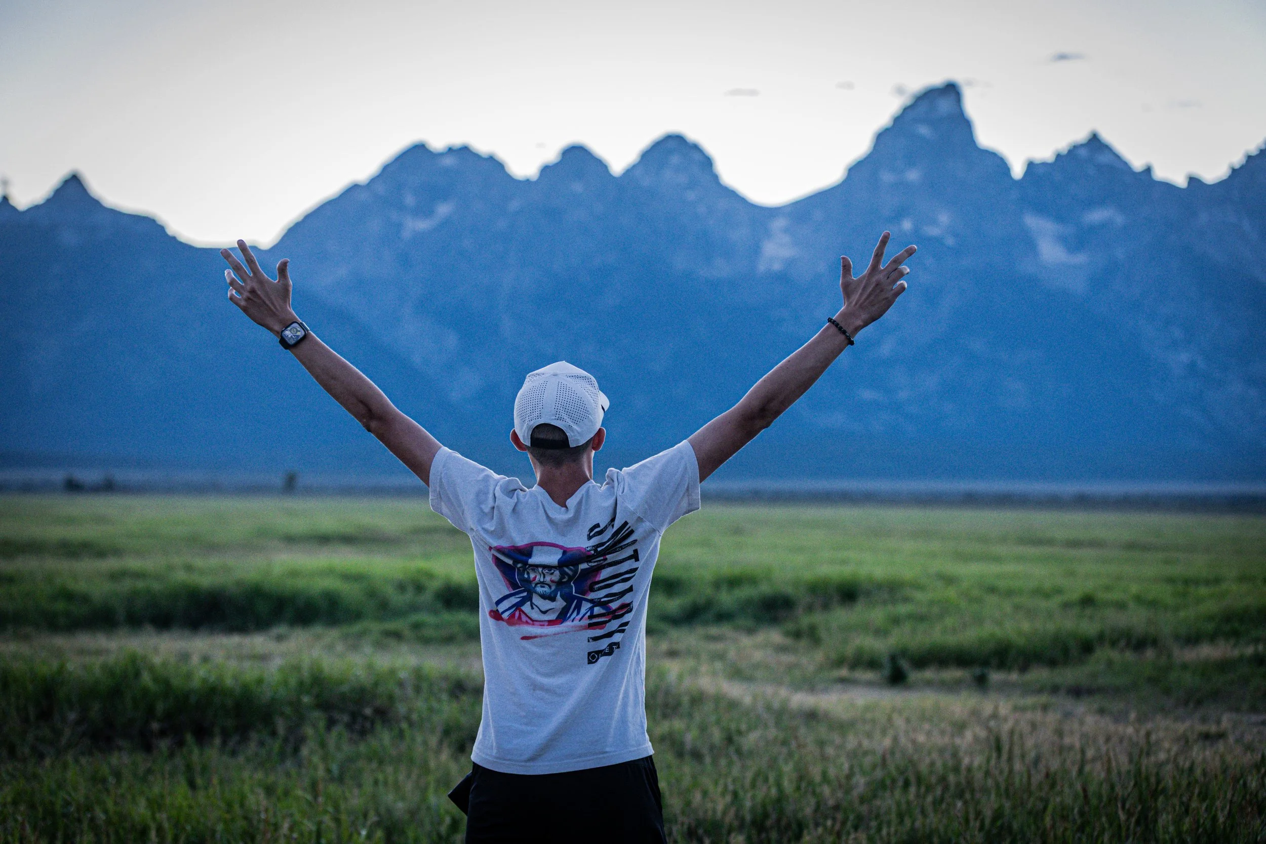 A person wearing a white cap and t-shirt with a graphic design, standing in a grassy field with arms raised, facing mountains in the distance at dusk or dawn.