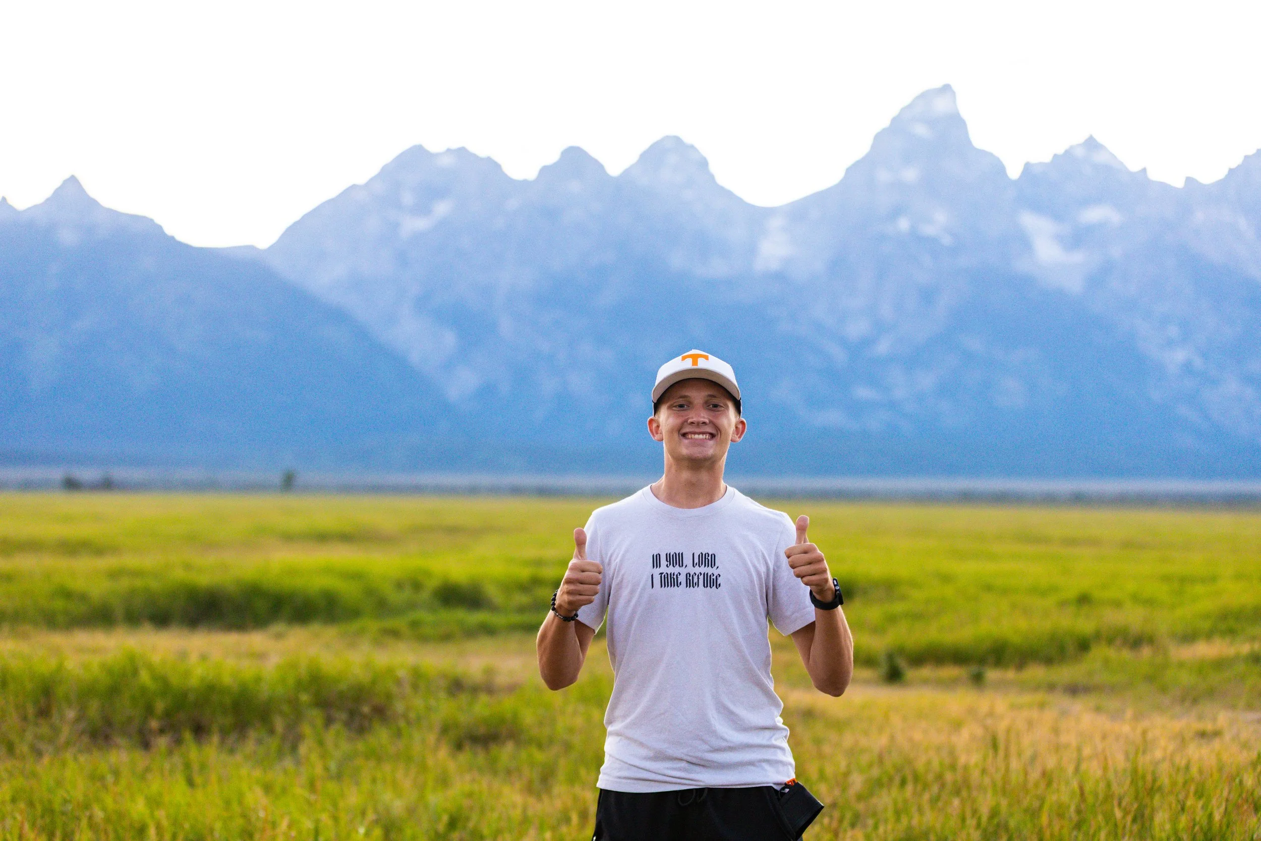 A young man in a white T-shirt and cap smiling and giving two thumbs up in a grassy field with mountains in the background.