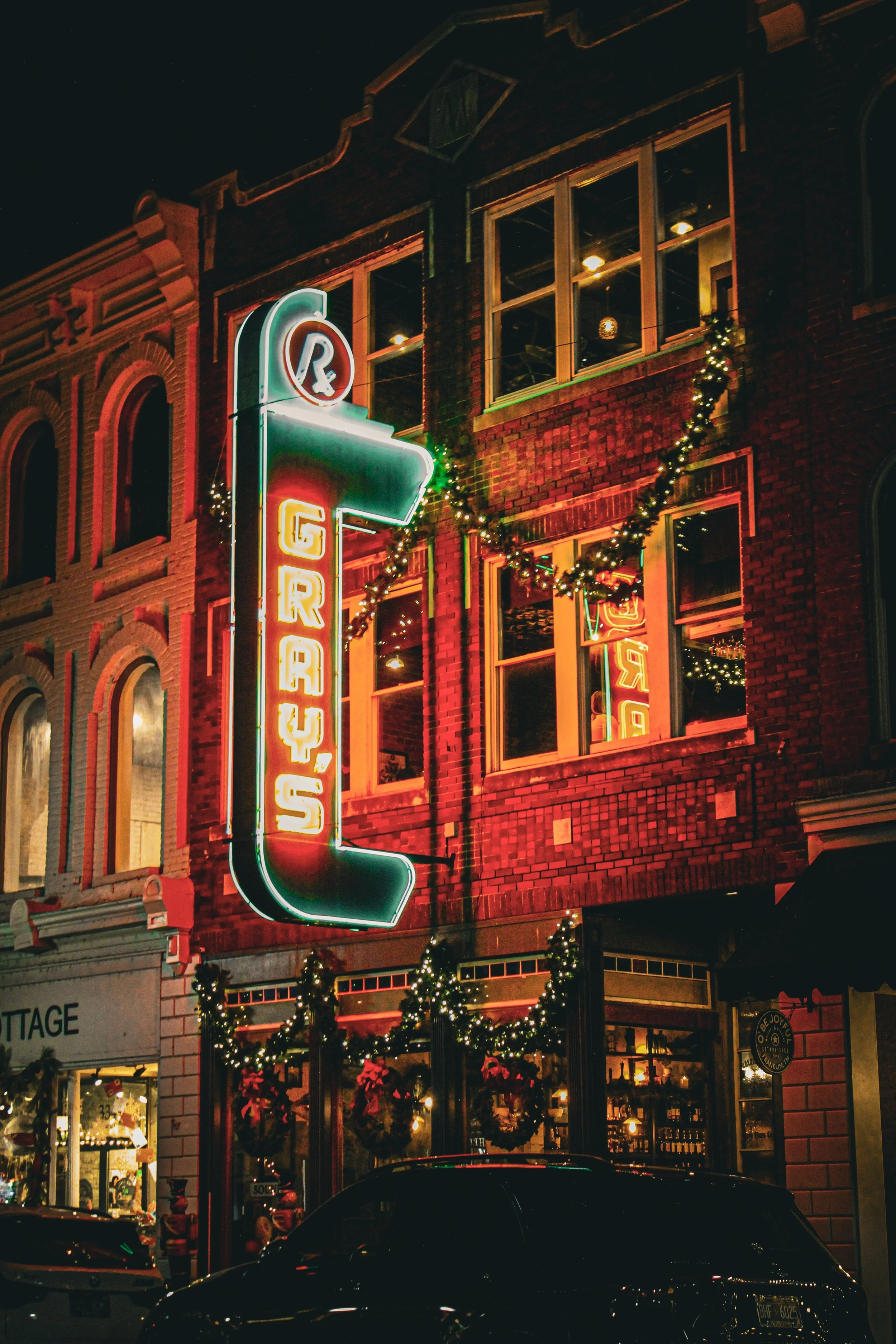 Night scene of a brick building decorated with Christmas lights and greenery. Neon signs display the words 'Bar' and 'Grocery.' The building has large windows showing interior lighting and decor.