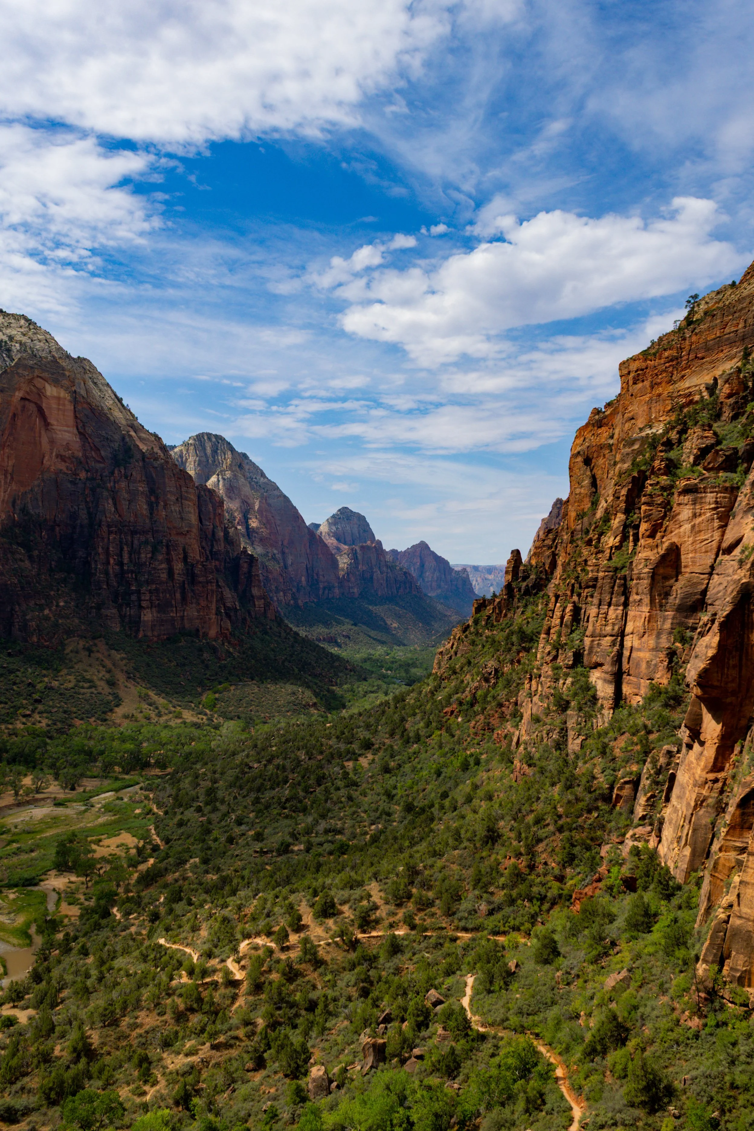 Scenic view of a canyon with steep, reddish-orange cliffs on both sides, green vegetation in the valley below, and a partly cloudy blue sky overhead.